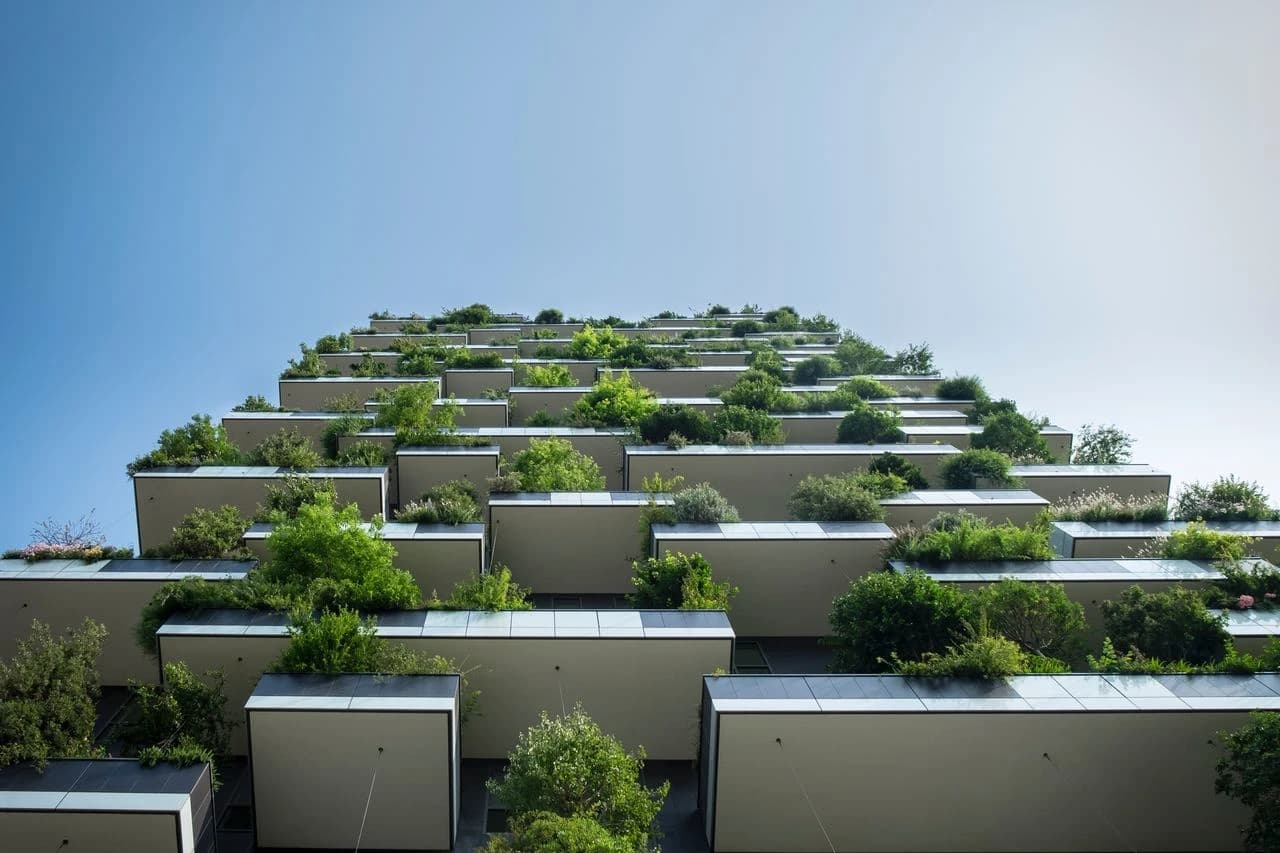 Façade d'un immeuble moderne avec des balcons étagés regorgeant de végétation luxuriante, formant une forêt verticale de plantes vertes et d'arbustes sous un ciel bleu clair.