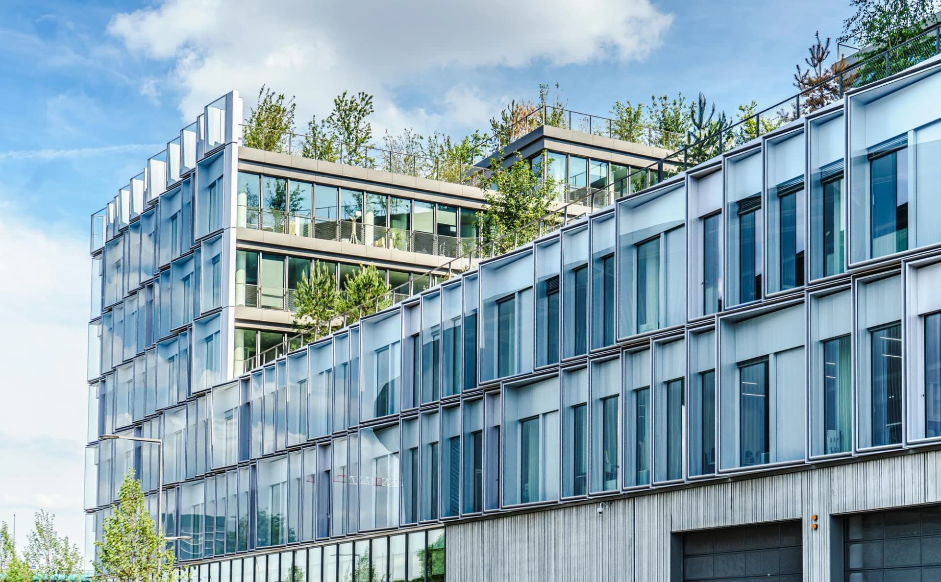 Bâtiment de bureaux moderne avec une façade de verre et des toits végétalisés luxuriants, reflétant le ciel bleu parsemé de nuages blancs.