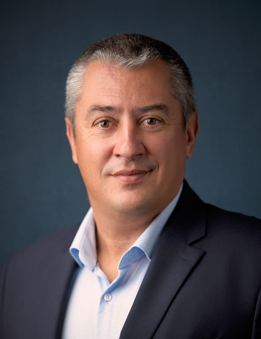 Portrait d'un homme d'âge moyen, cheveux gris, souriant légèrement, portant un costume bleu marine et une chemise bleue claire, sur fond bleu foncé.