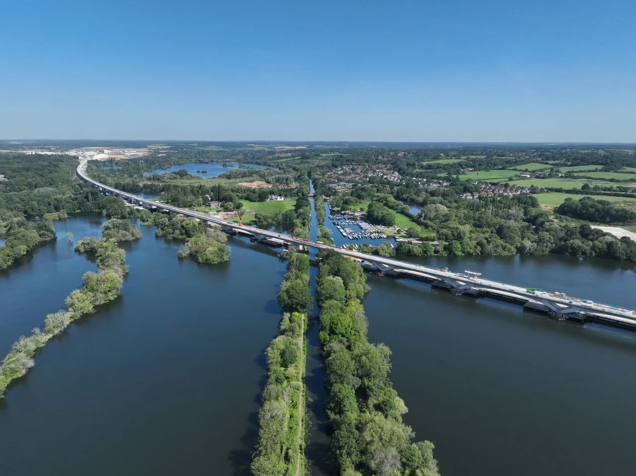 Vue aérienne d'un long pont routier moderne en construction enjambant une vaste étendue d'eau foncée parsemée d'îles boisées. Le paysage environnant est verdoyant avec des zones rurales et une marina, sous un ciel bleu clair.