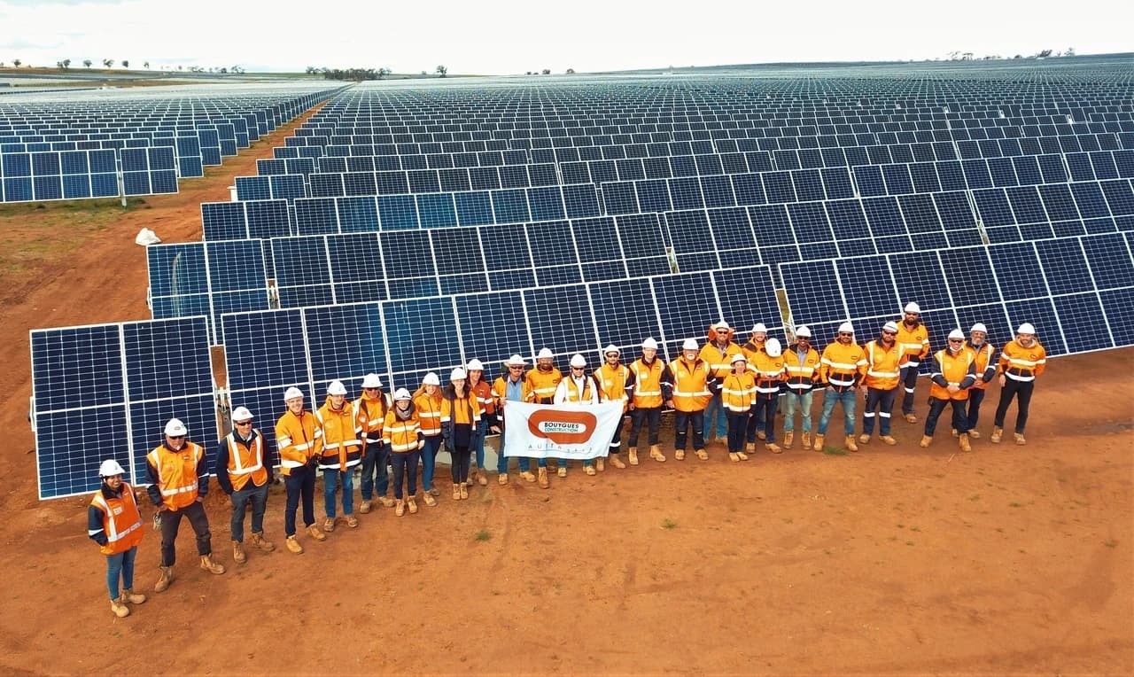Un grand groupe de travailleurs en gilets orange haute visibilité et casques blancs se tient sur un sol ocre, tenant une bannière "Bouygues Construction Australia", devant un vaste champ de panneaux solaires bleus s'étendant à l'horizon sous un ciel lumineux.