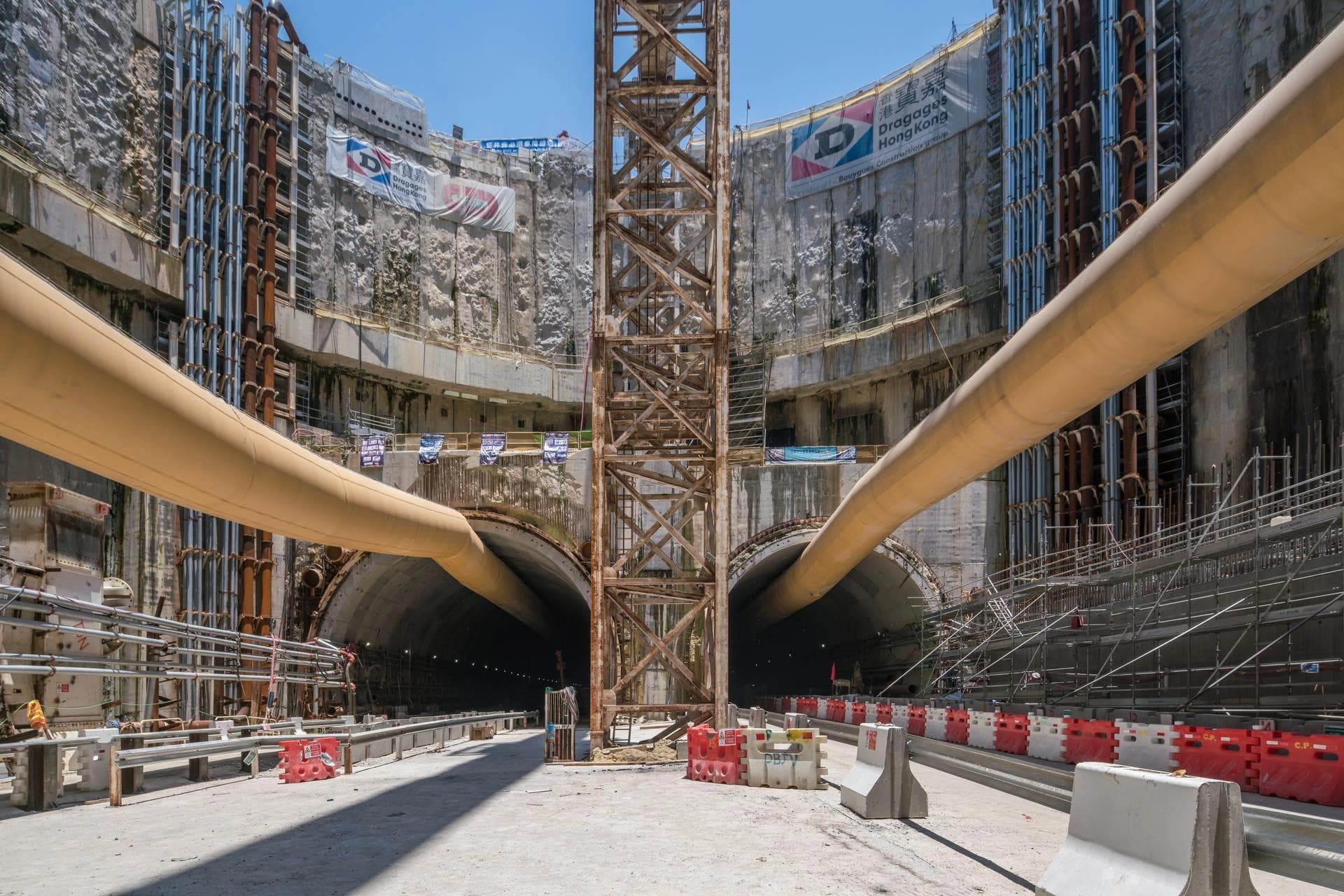 Une excavation massive révèle deux grandes entrées de tunnel sombres en construction, encadrées par des murs de béton brut, des échafaudages rouillés et des bannières Dragages Hong Kong. Des tuyaux jaunes proéminents traversent la scène, avec une structure métallique rouillée et des barrières de sécurité rouges et blanches au premier plan, sous un ciel bleu vif.