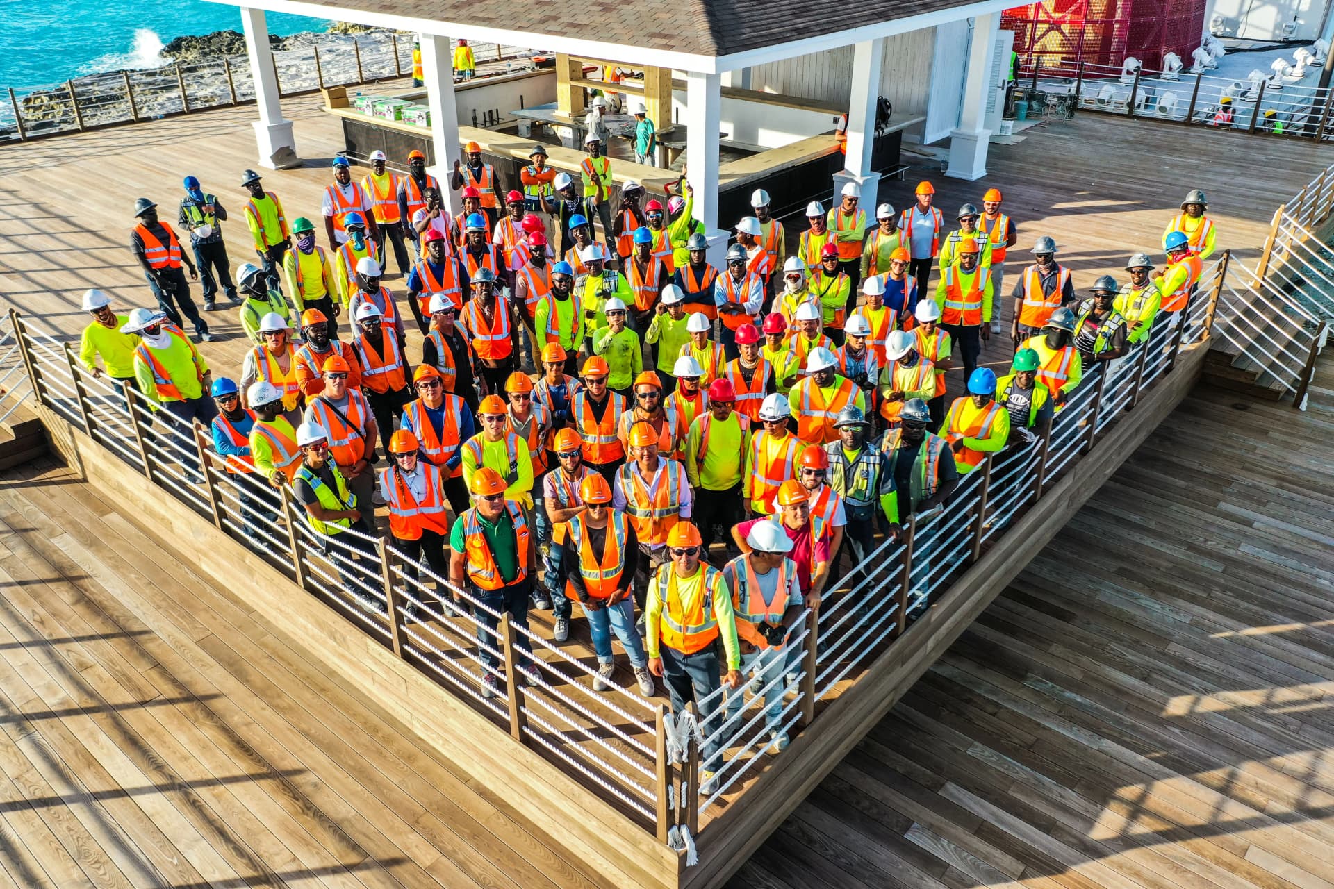 Un groupe important d'ouvriers du bâtiment, coiffés de casques de chantier de différentes couleurs et vêtus de gilets haute visibilité orange vif ou vert citron, posent pour une photo sur une grande terrasse en bois surplombant une structure de bar et l'océan bleu.
