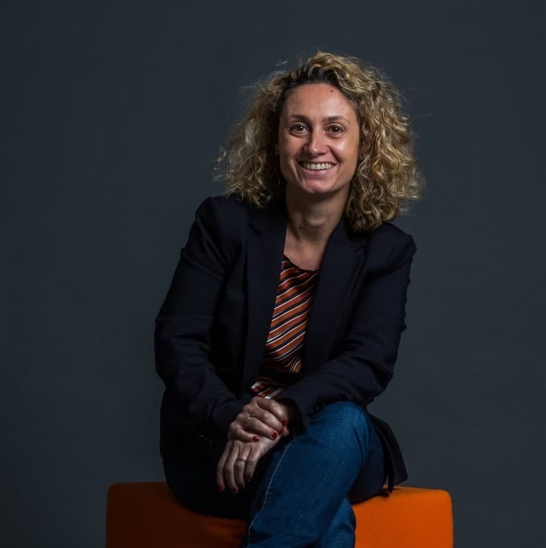 Femme souriante aux cheveux blonds bouclés, vêtue d'un blazer foncé et d'un jean, assise sur un tabouret orange.