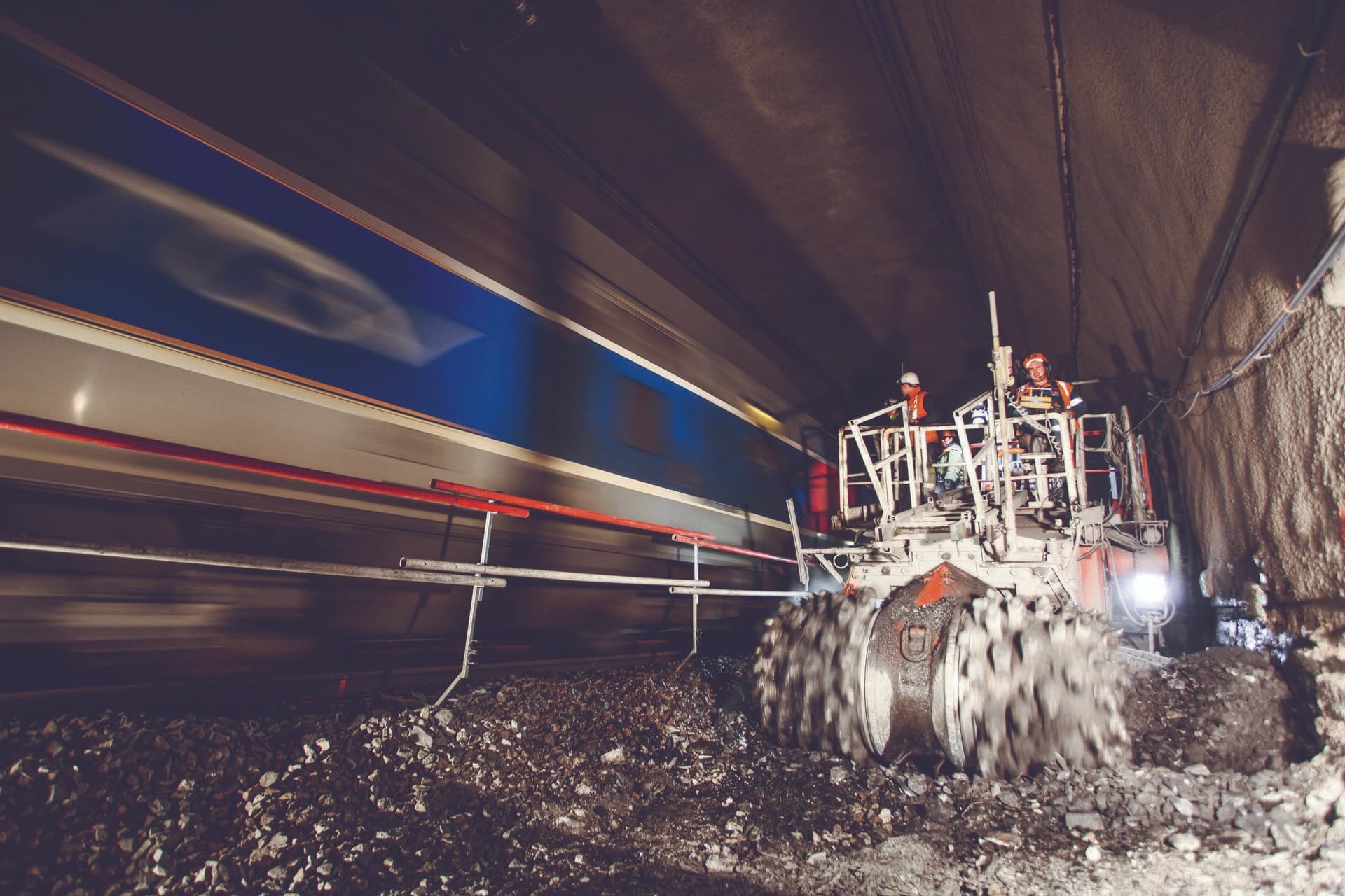 Des ouvriers en gilets orange sur une machine de chantier avec un tambour rotatif soulèvent du ballast et de la terre, tandis qu'un train bleu et gris passe à toute vitesse dans un tunnel éclairé.