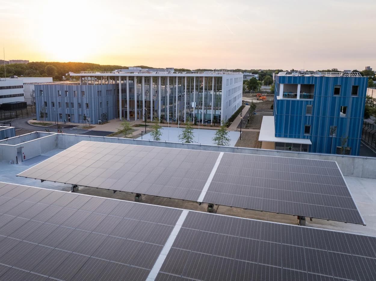 Vue aérienne d'un complexe moderne avec de vastes panneaux solaires en premier plan, des bâtiments gris, blancs et un bâtiment bleu vif à l'arrière-plan, sous un ciel de coucher de soleil doré.