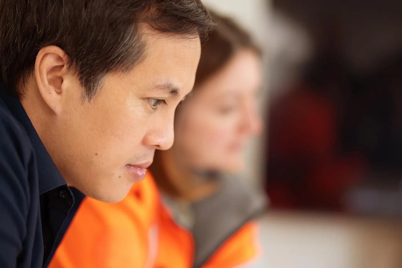 Gros plan en profil d'un homme aux cheveux foncés portant une chemise bleu marine, regardant attentivement vers le bas. Une personne vêtue d'orange est floue en arrière-plan, suggérant une collaboration.