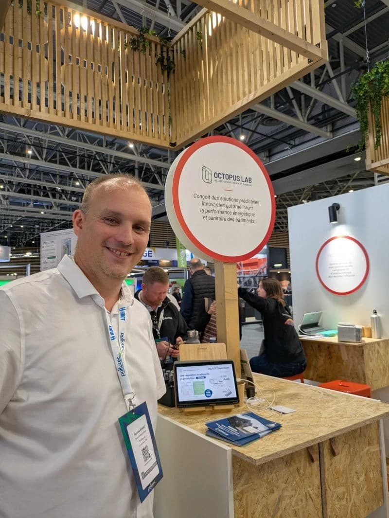 Un homme souriant en chemise blanche se tient devant un stand d'exposition "OCTOPUS LAB", dont le panneau blanc et rouge mentionne des solutions pour améliorer la performance énergétique des bâtiments, avec une tablette et un comptoir en bois.