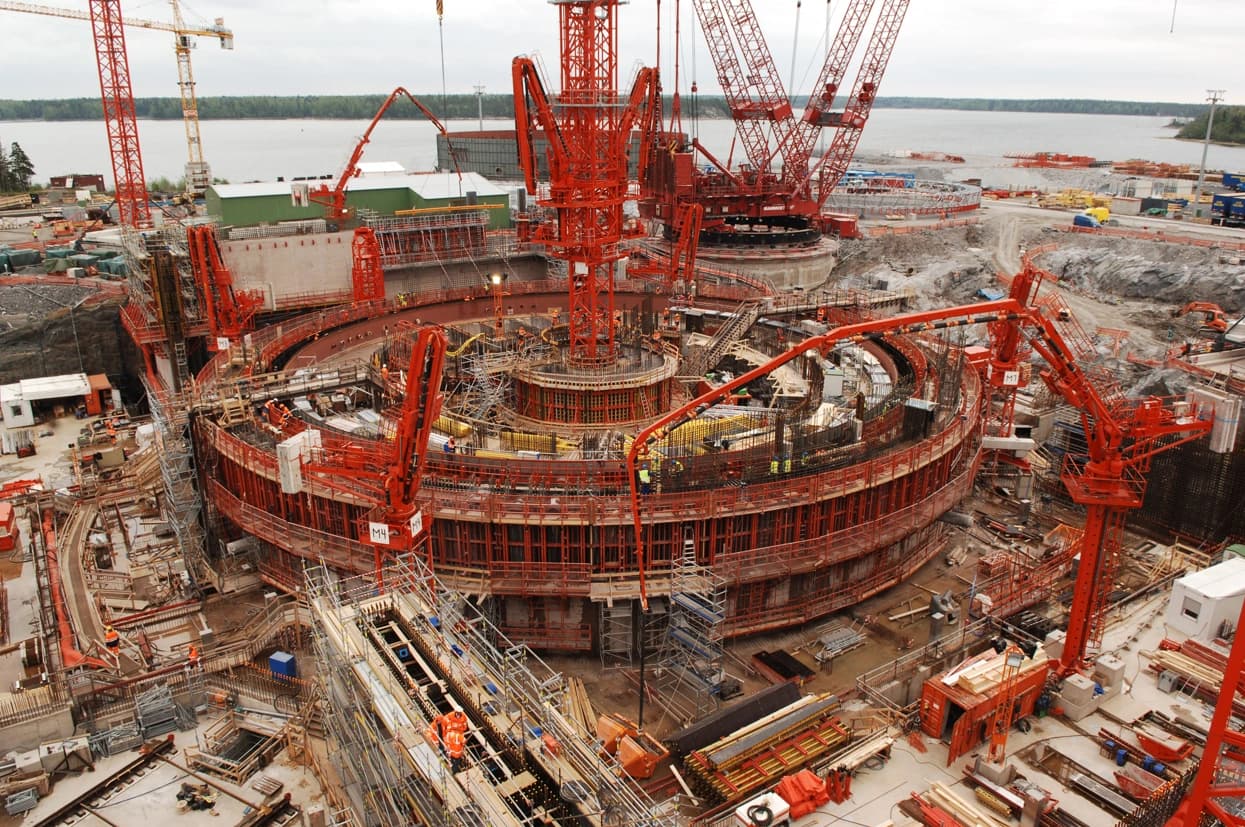 Vue panoramique d'un immense chantier de construction pour une structure circulaire, dominé par de nombreuses pompes à béton et grues rouges intenses, avec des travailleurs s'affairant parmi les échafaudages et le ferraillage sous un ciel couvert, près d'un plan d'eau.
