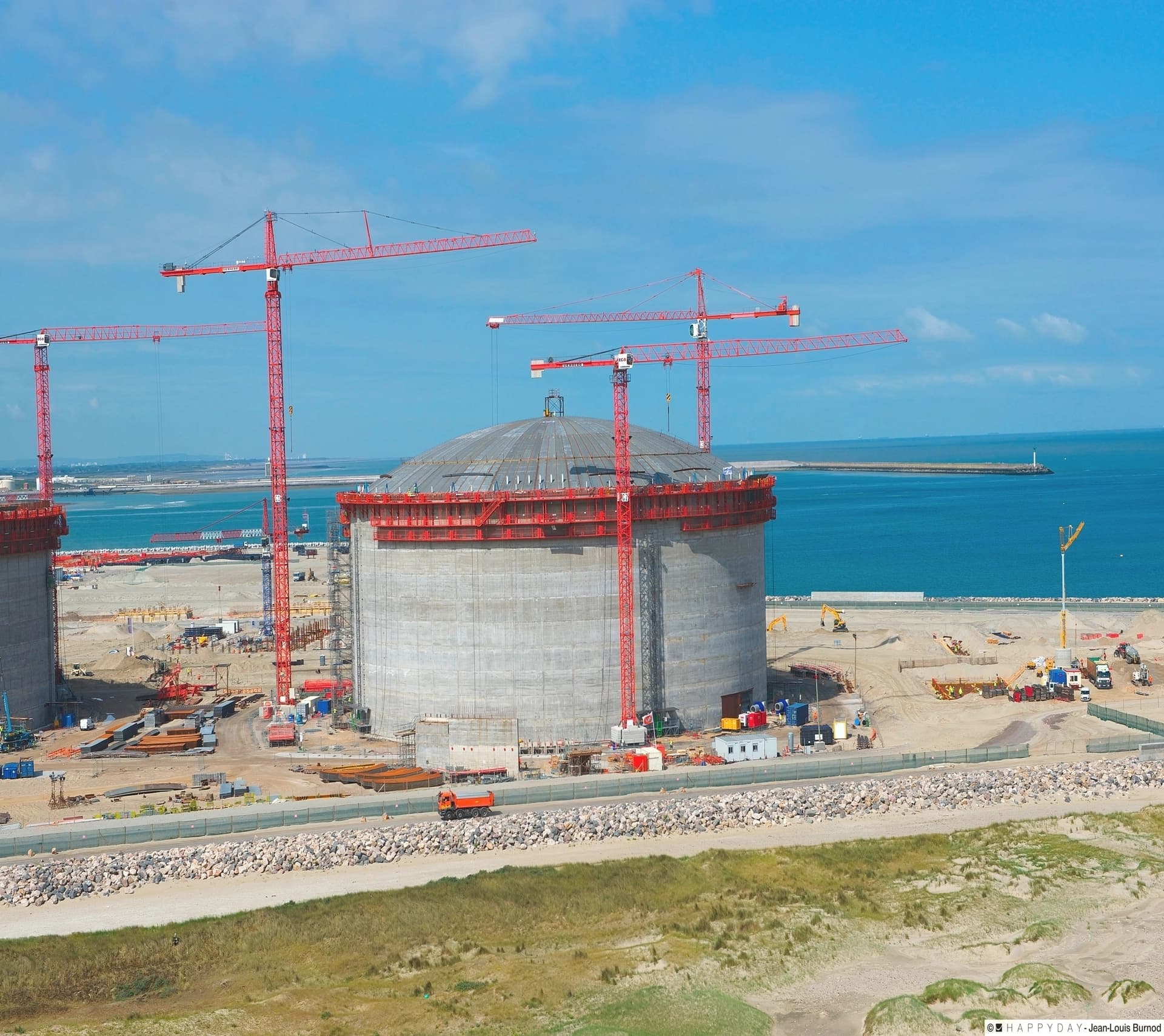 Vue aérienne d'un chantier de construction côtier montrant de grands réservoirs de stockage en béton gris, l'un coiffé d'un dôme, entourés de plusieurs grues rouges sous un ciel bleu clair, avec la mer turquoise en arrière-plan.