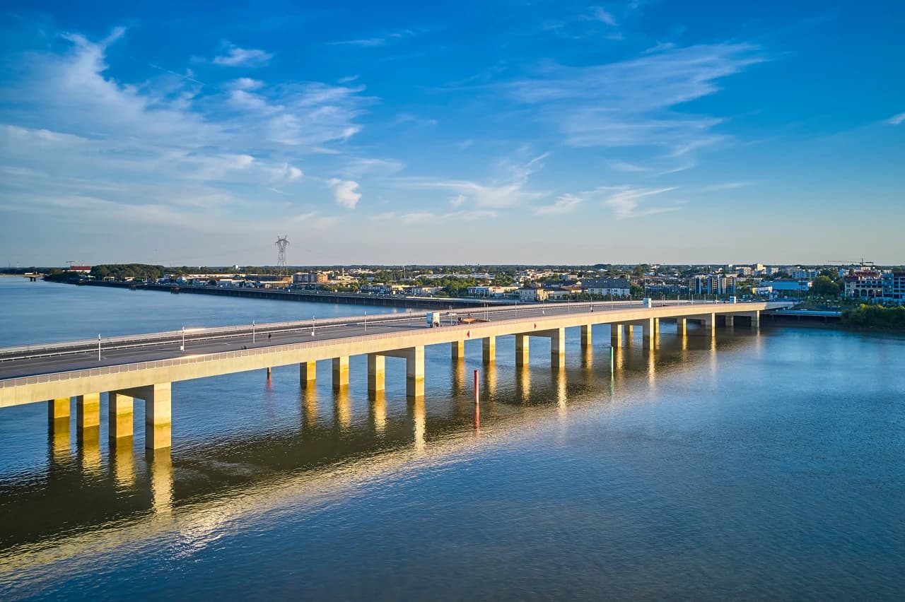 Vue aérienne d'un long pont en béton traversant une rivière, avec des reflets dorés du soleil sur l'eau et une ville lointaine en arrière-plan sous un ciel bleu parsemé de nuages.