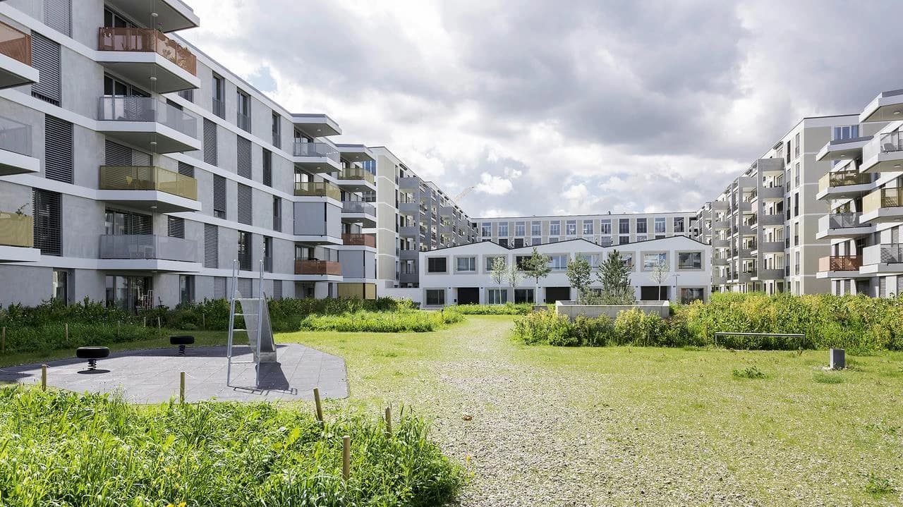 Modern light grey apartment buildings with balconies featuring varied colored railings surround a vibrant green courtyard containing a playground, all under a cloudy sky.