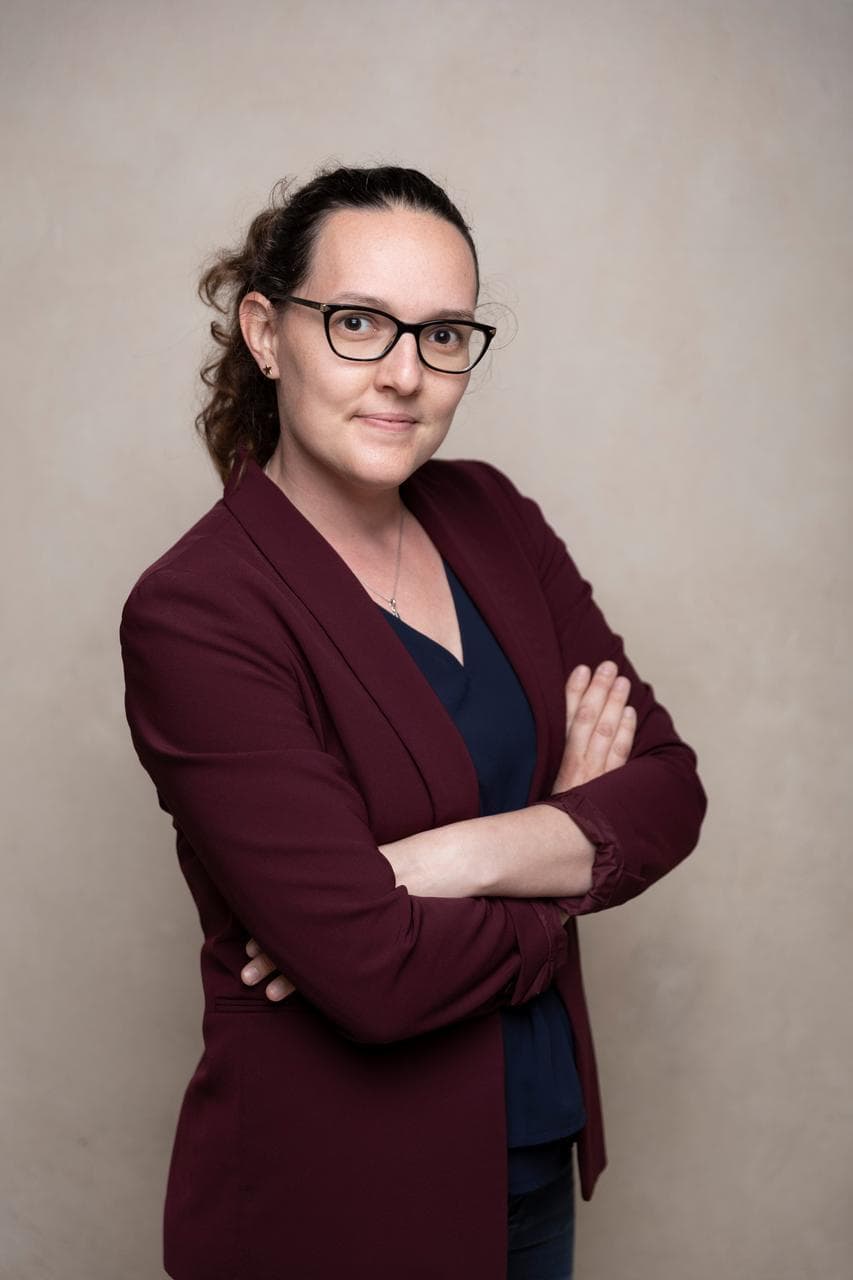 Portrait d'une femme aux cheveux foncés et lunettes, portant un blazer bordeaux et un haut bleu, les bras croisés, avec un léger sourire.