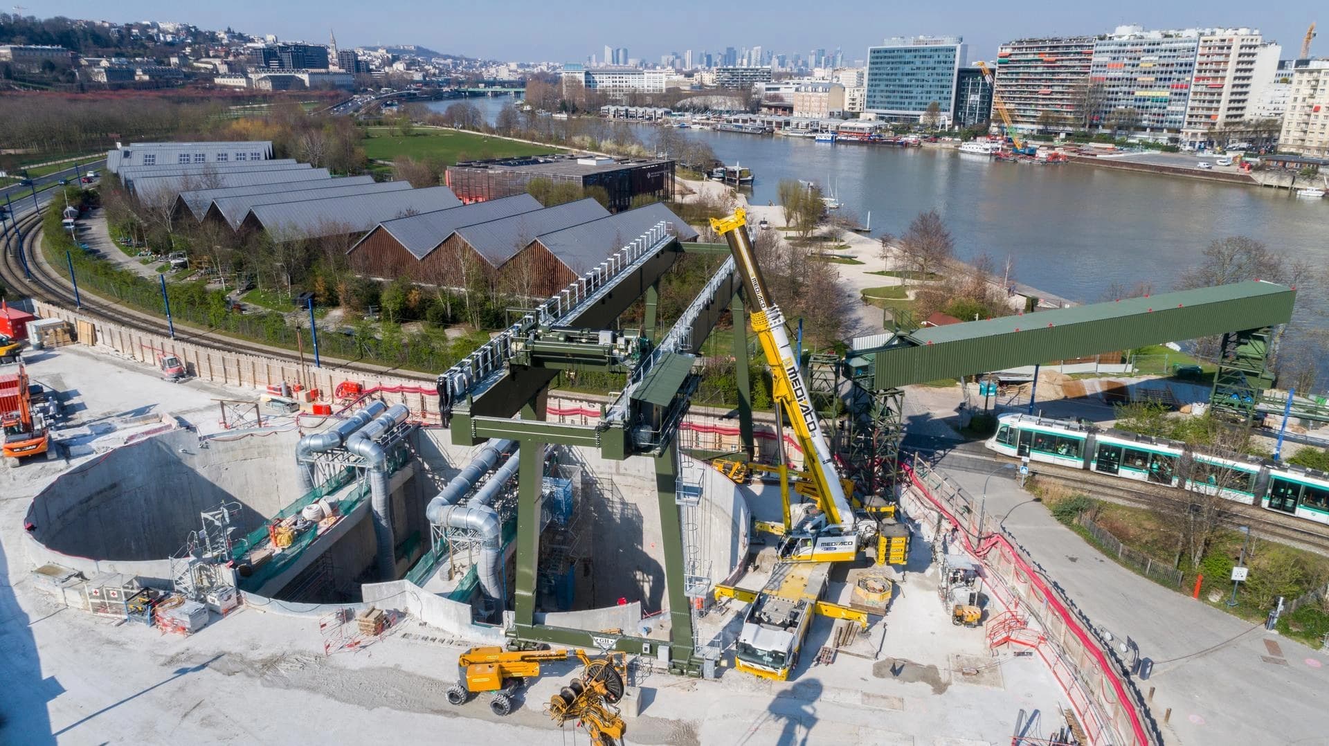Vue aérienne d'un grand chantier de construction urbain avec une fosse circulaire profonde, une grue portique verte et une grue mobile jaune. Un tramway circule à côté, avec une rivière et des bâtiments urbains en arrière-plan sous un ciel clair.