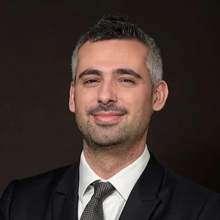 Portrait d'un homme souriant aux cheveux courts grisonnants et barbe foncée, portant un costume sombre et une cravate grise sur fond uni.