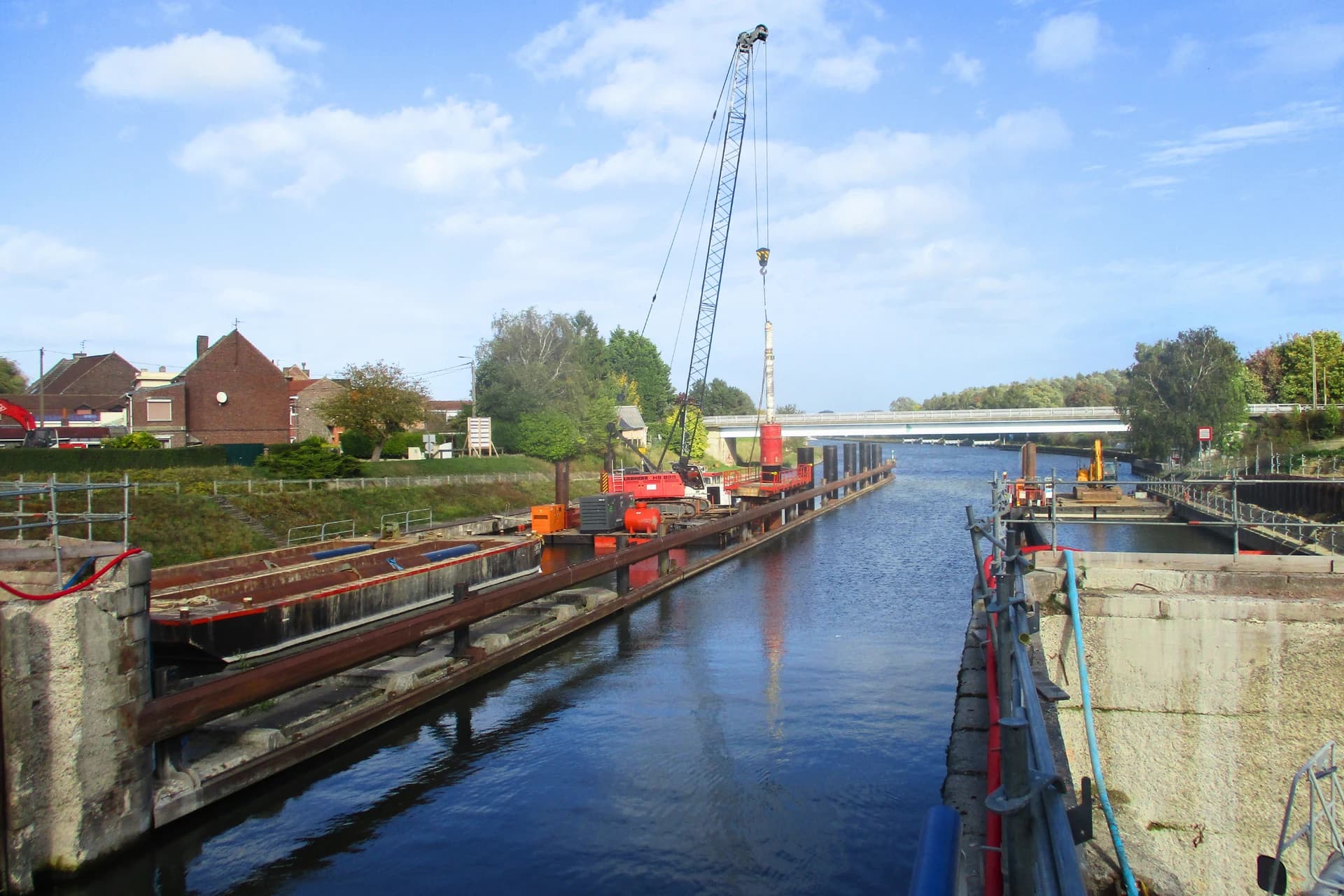 Un grand pont roulant rouge sur une barge est visible dans un large canal, soulevant un élément sous un ciel bleu clair. Des maisons résidentielles bordent la rive gauche tandis qu'un pont blanc enjambe la voie navigable au loin.