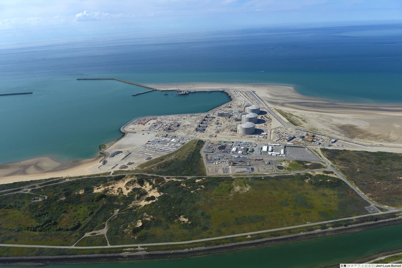 Vue aérienne d'un terminal GNL industriel avec de grands réservoirs de stockage gris et un port côtier sur la mer bleue. Le complexe est entouré de plages de sable, de végétation et d'un canal en premier plan.