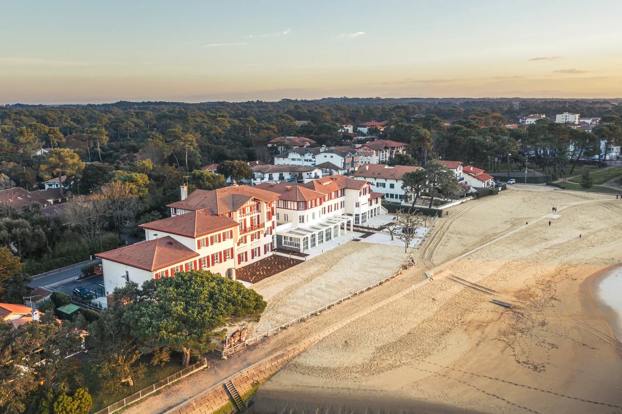 Vue aérienne d'un élégant complexe hôtelier blanc aux toits et volets rouges, niché entre une large plage de sable doré et une forêt de pins luxuriante, sous la douce lumière rasante du soleil.