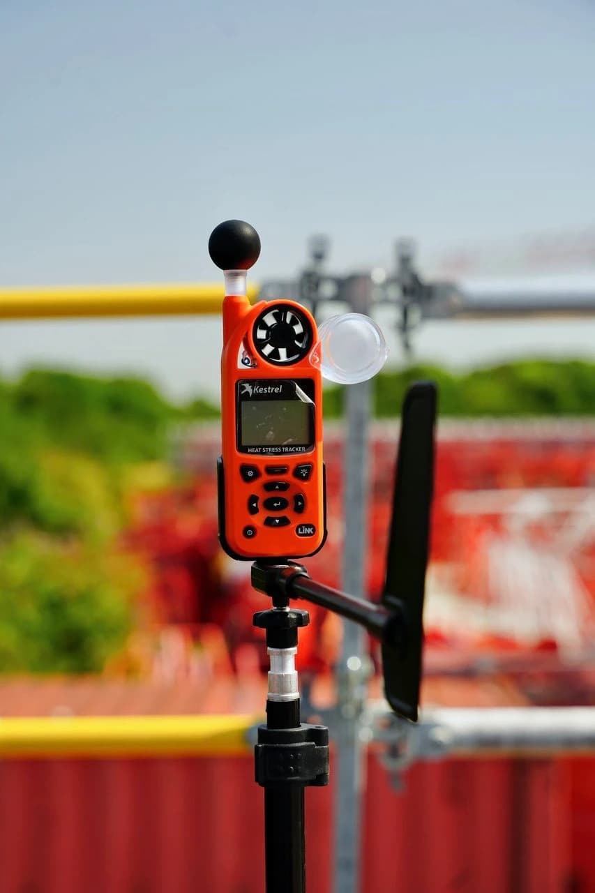 Un appareil Kestrel Heat Stress Tracker orange, doté d'un capteur à boule noire et d'un petit ventilateur, est monté sur un trépied noir en extérieur. L'arrière-plan flou révèle un ciel bleu clair, une balustrade jaune et des structures rouges, suggérant un environnement de chantier ou industriel.