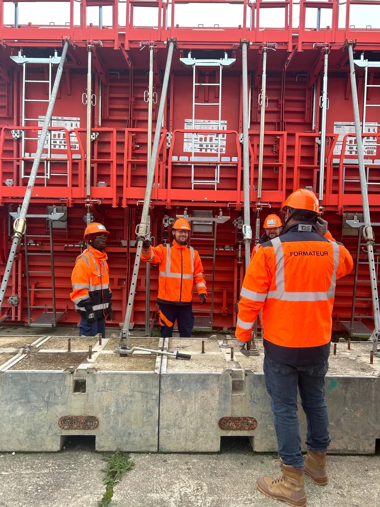 Quatre ouvriers en gilets de haute visibilité orange et casques de sécurité devant des panneaux de coffrage rouges. L'un d'eux a "FORMATEUR" écrit dans le dos, indiquant une session de formation ou d'instruction.