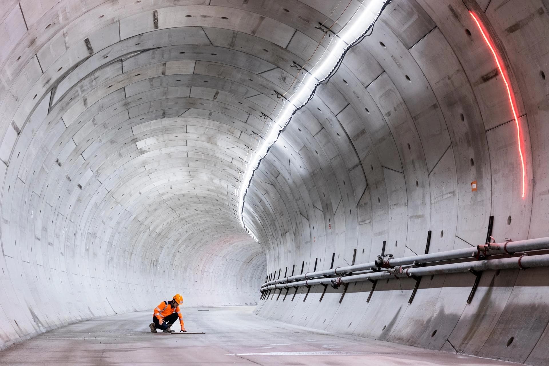 Un travailleur en gilet orange fluo est accroupi sur le sol en béton gris d'un vaste tunnel nouvellement construit, éclairé par une bande lumineuse blanche brillante d'un côté et un néon rouge de l'autre. Des tuyaux gris courent le long du mur droit inférieur du tunnel courbé.
