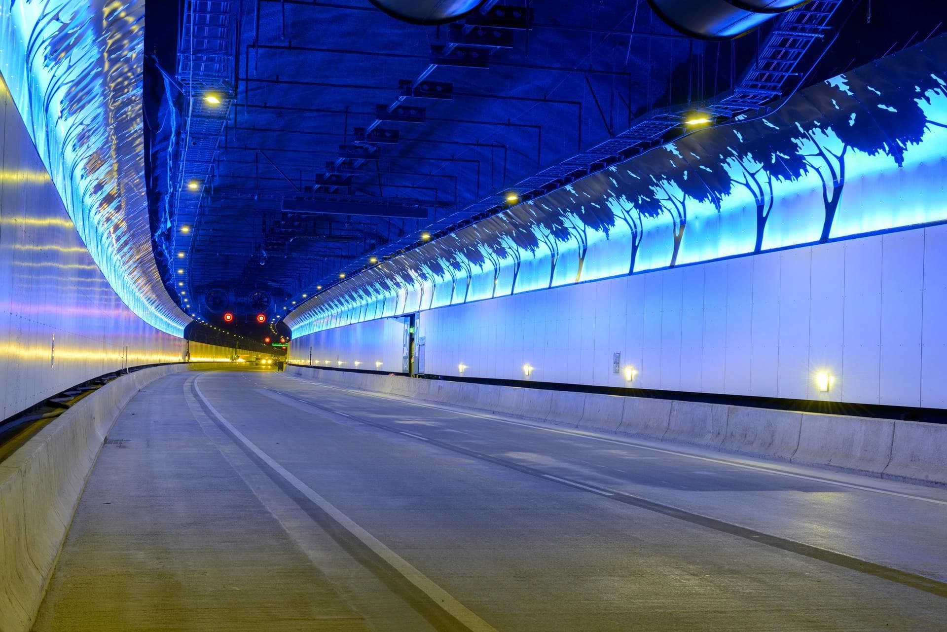 Tunnel routier moderne et lumineux, éclairé principalement en bleu avec des silhouettes d'arbres stylisées sur la droite et des reflets jaunes sur la gauche, tandis qu'une route vide à deux voies s'y courbe.