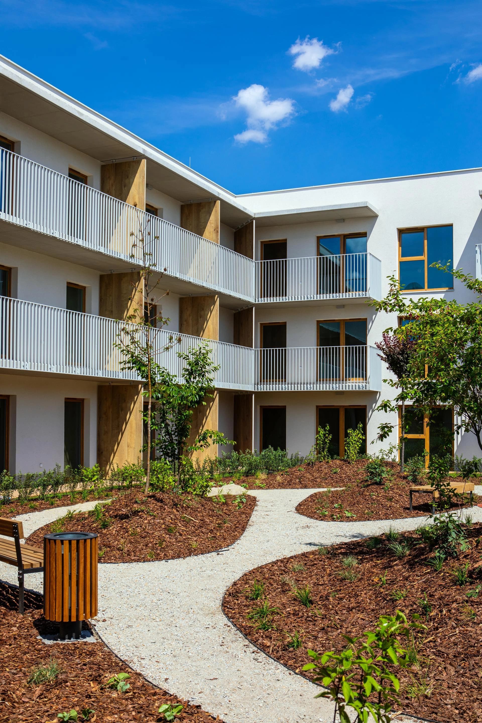 Immeuble moderne avec balcons blancs et une cour paysagée dotée de sentiers de gravier et de bancs.