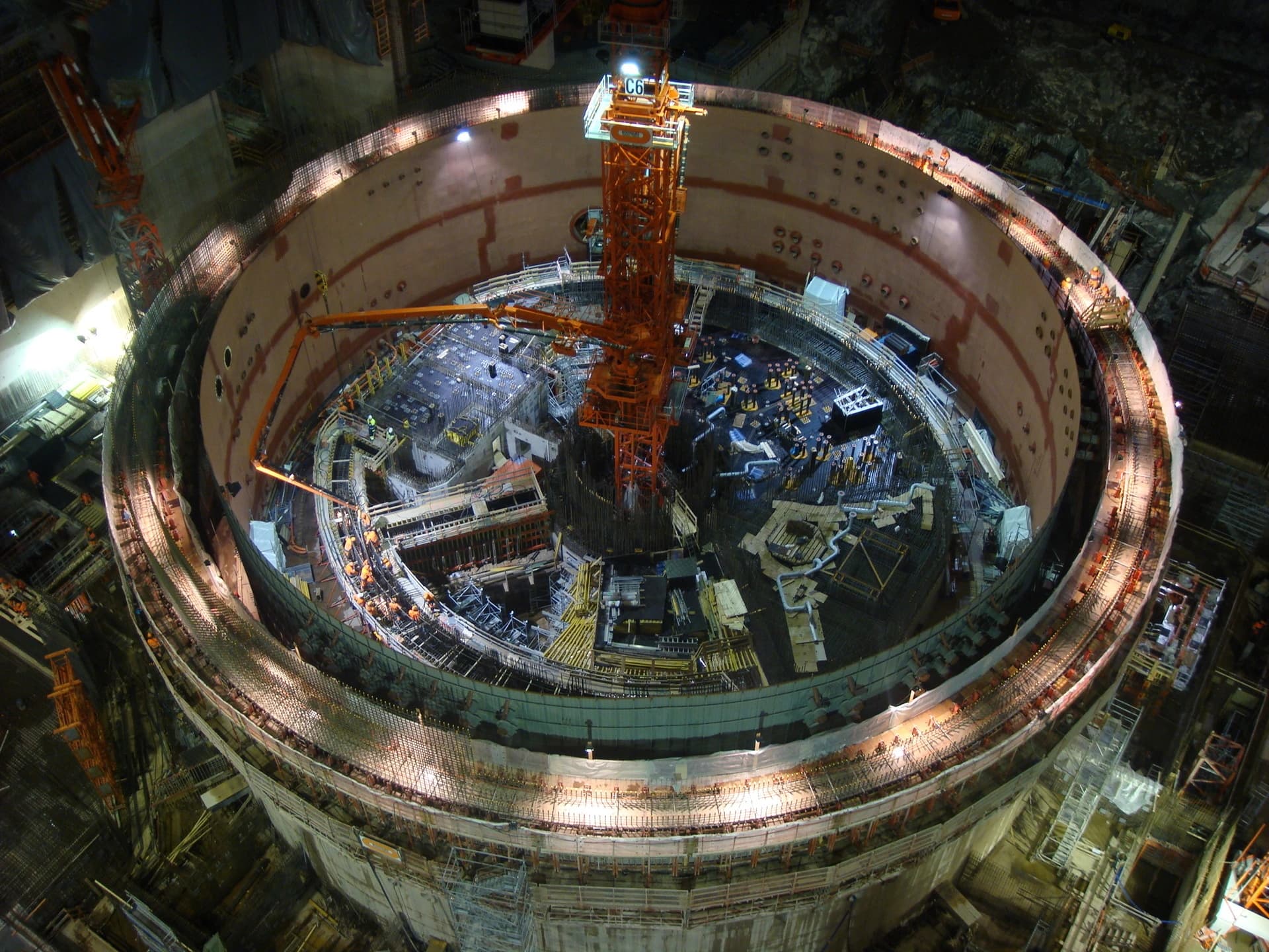 Vue aérienne nocturne d'un gigantesque chantier de construction circulaire avec une enceinte en béton beige. Un grand mât de grue orange, des pompes à béton et de nombreux ouvriers s'activent sous des projecteurs, révélant des échafaudages et des renforcements métalliques.