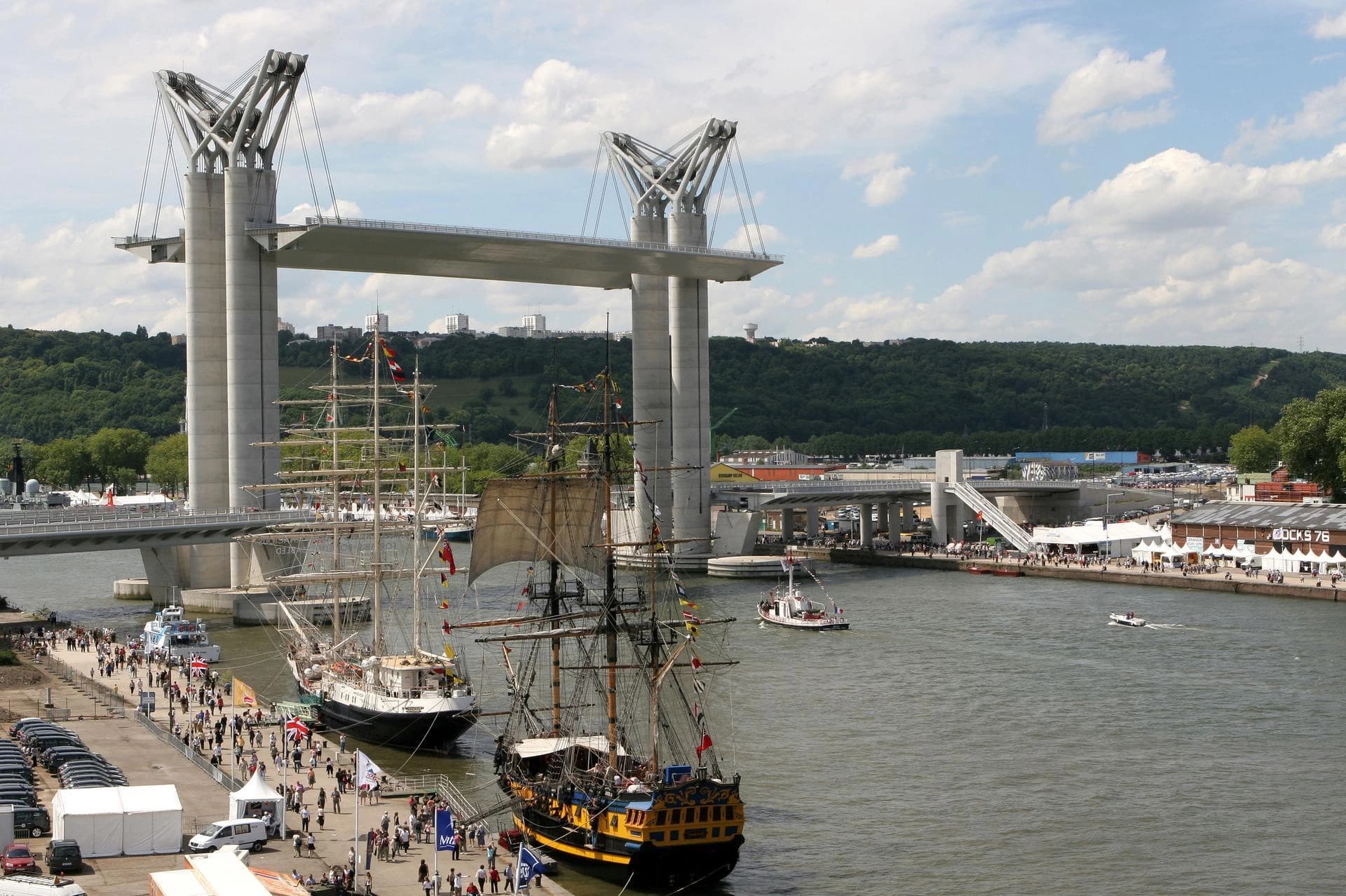 Deux grands voiliers, l'un noir et blanc, l'autre noir et jaune, sont amarrés à un quai animé rempli de spectateurs, sous un pont haubané moderne gris et des collines verdoyantes sur la rive opposée.