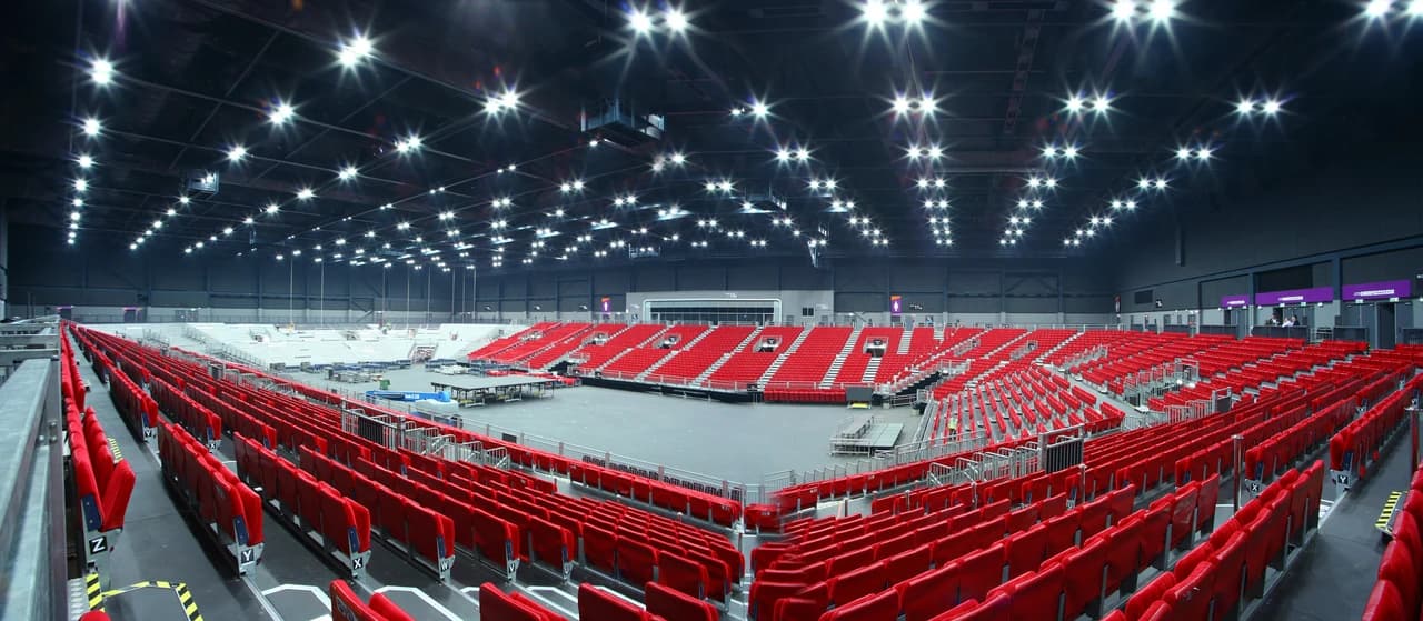 Vue panoramique d'une grande arène intérieure moderne, avec des rangées de sièges rouges vides entourant un vaste espace central sous un plafond sombre illuminé par de nombreuses lumières vives.