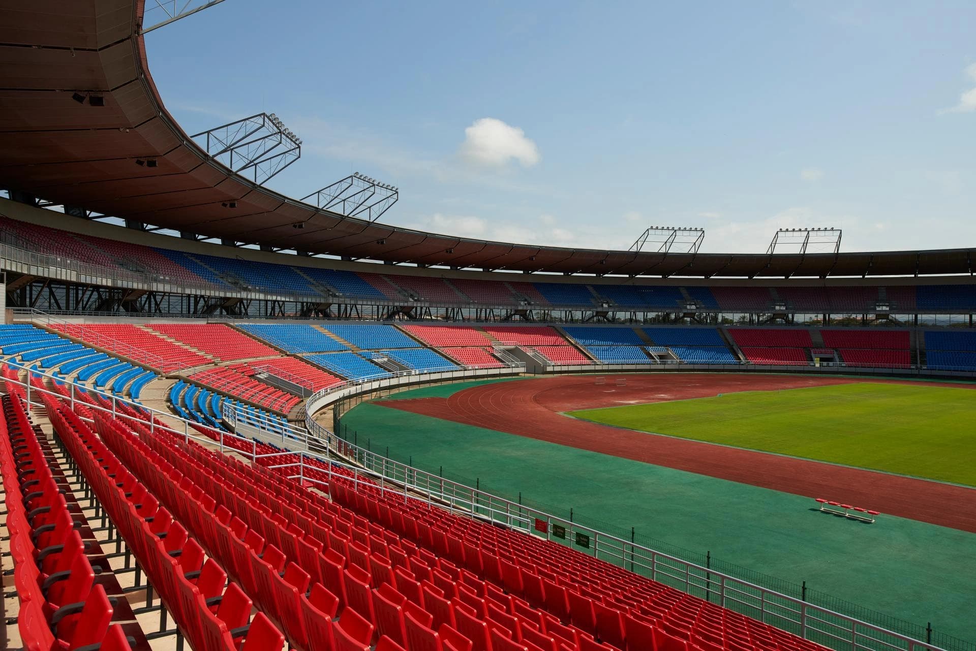 Un grand stade de sport vide avec des sièges rouges et bleus, une piste d'athlétisme et un terrain vert sous un ciel bleu.