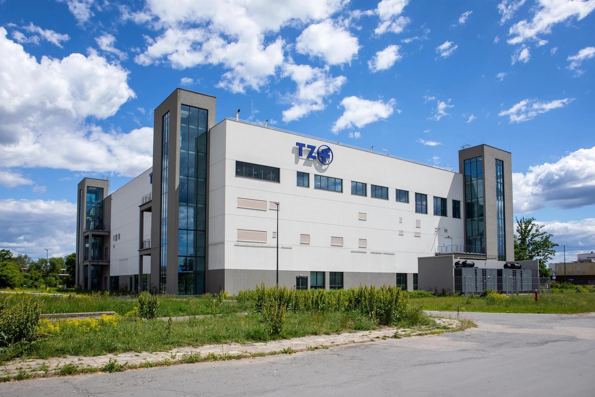 Un bâtiment industriel blanc moderne avec un logo TZO bleu sous un ciel bleu clair parsemé de nuages.