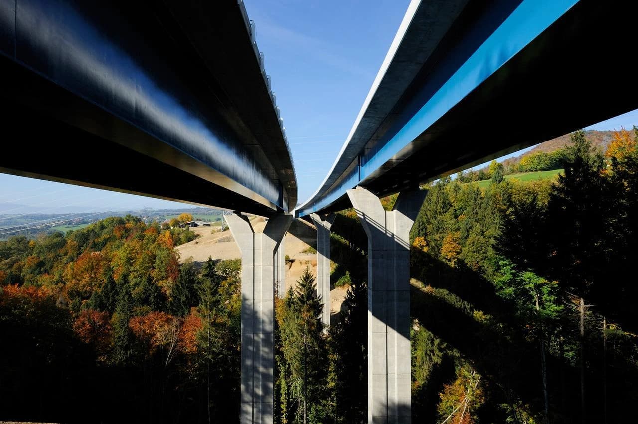 Deux viaducs parallèles sombres, dont l'un arbore une bande bleue distinctive, traversent une vallée boisée aux couleurs automnales sous un ciel bleu clair. De hauts piliers de béton soutiennent les ponts, projetant de longues ombres sur la forêt.