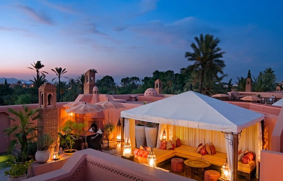 Une luxueuse terrasse de toit de style marocain au crépuscule, avec des bâtiments roses, des zones de détente illuminées sous des auvents blancs, des lanternes décoratives, des palmiers et des jardins luxuriants sous un ciel bleu-violet.