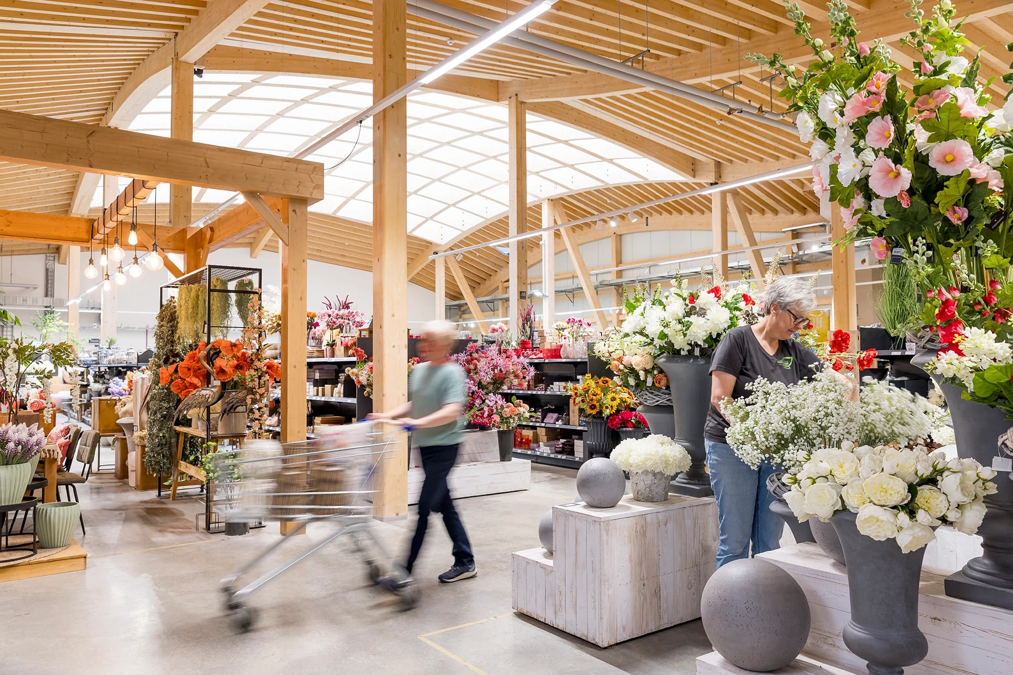 Un magasin de fleurs et de décoration moderne et lumineux, doté d'un haut plafond en bois avec des poutres et de larges puits de lumière. Une personne arrange des fleurs artificielles blanches tandis qu'un client, flou par le mouvement, pousse un chariot à travers les allées remplies de diverses fleurs colorées.
