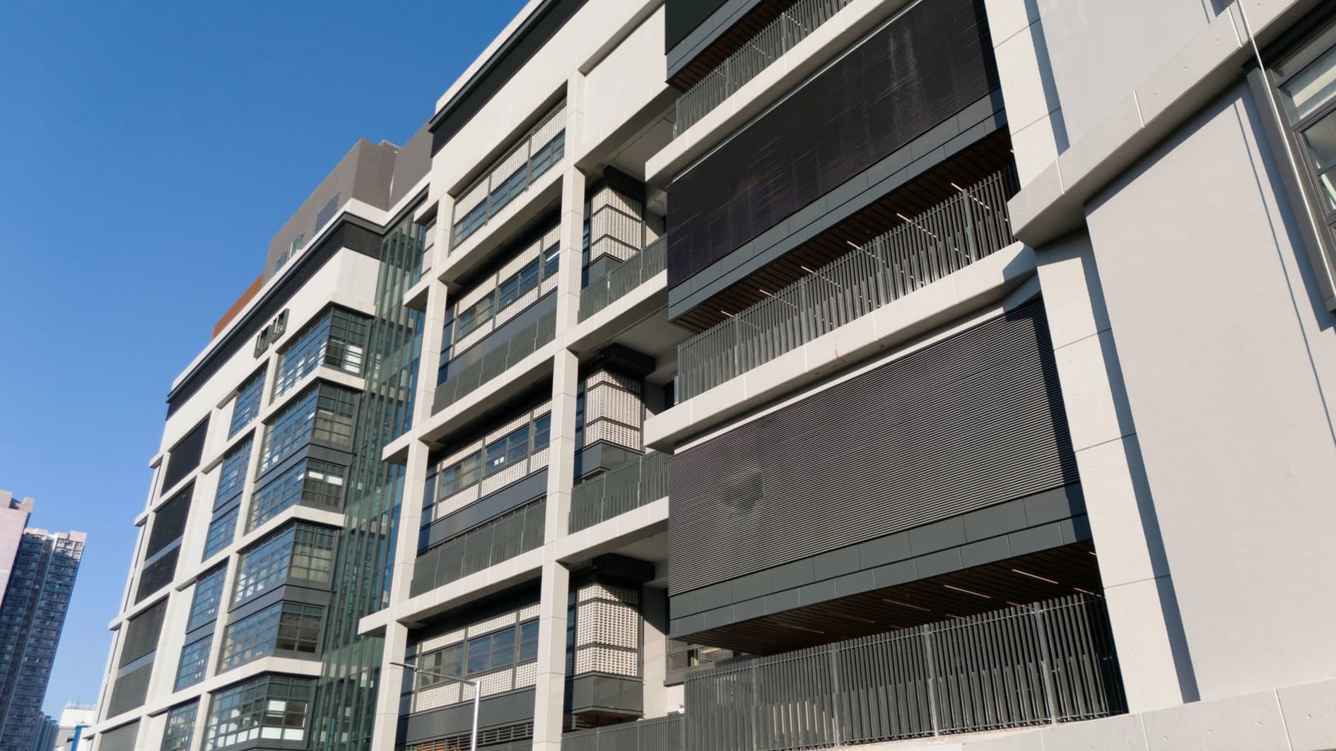 Façade moderne d'un bâtiment aux lignes épurées, avec des sections en béton clair contrastant avec de grandes fenêtres sombres et des panneaux gris foncé, sous un ciel bleu clair.