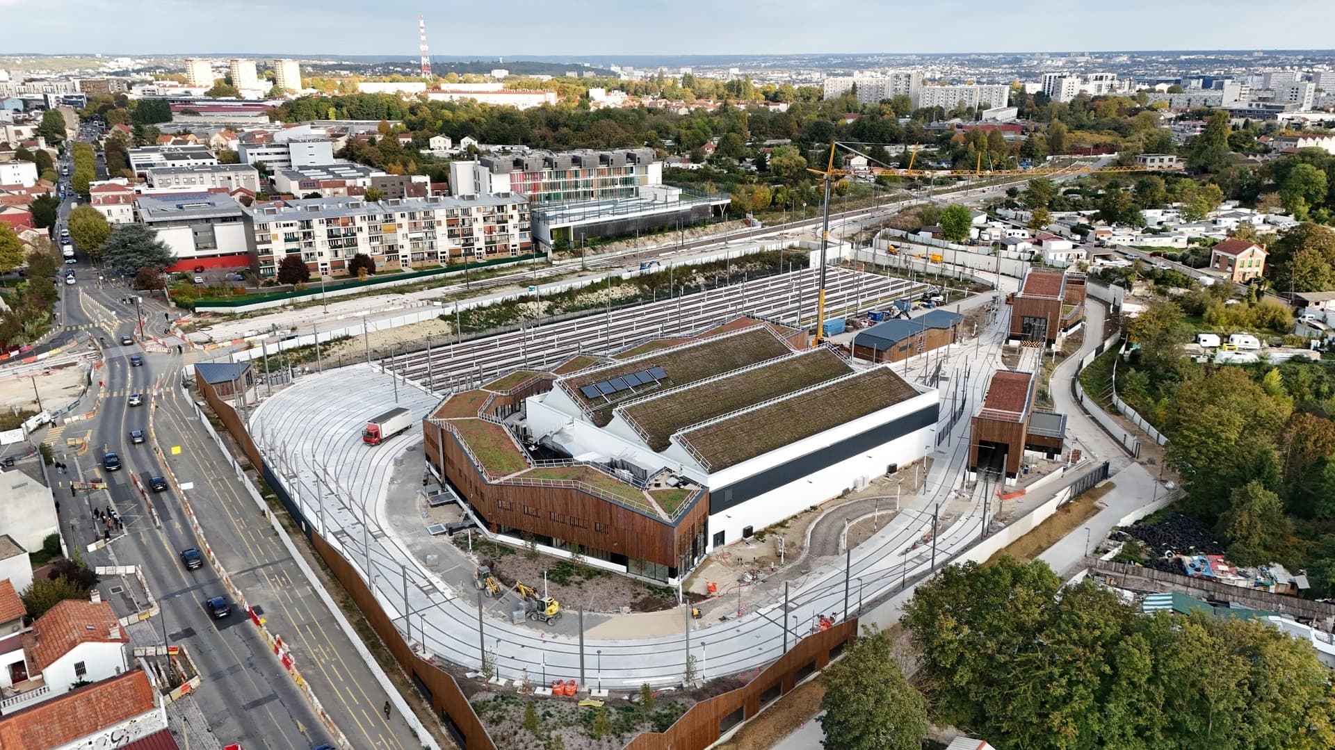 Vue aérienne d'un vaste dépôt de tramway moderne, doté de toits végétalisés avec panneaux solaires et de façades en bois, s'intégrant à un paysage urbain dense comprenant voies de tram, route et habitations.