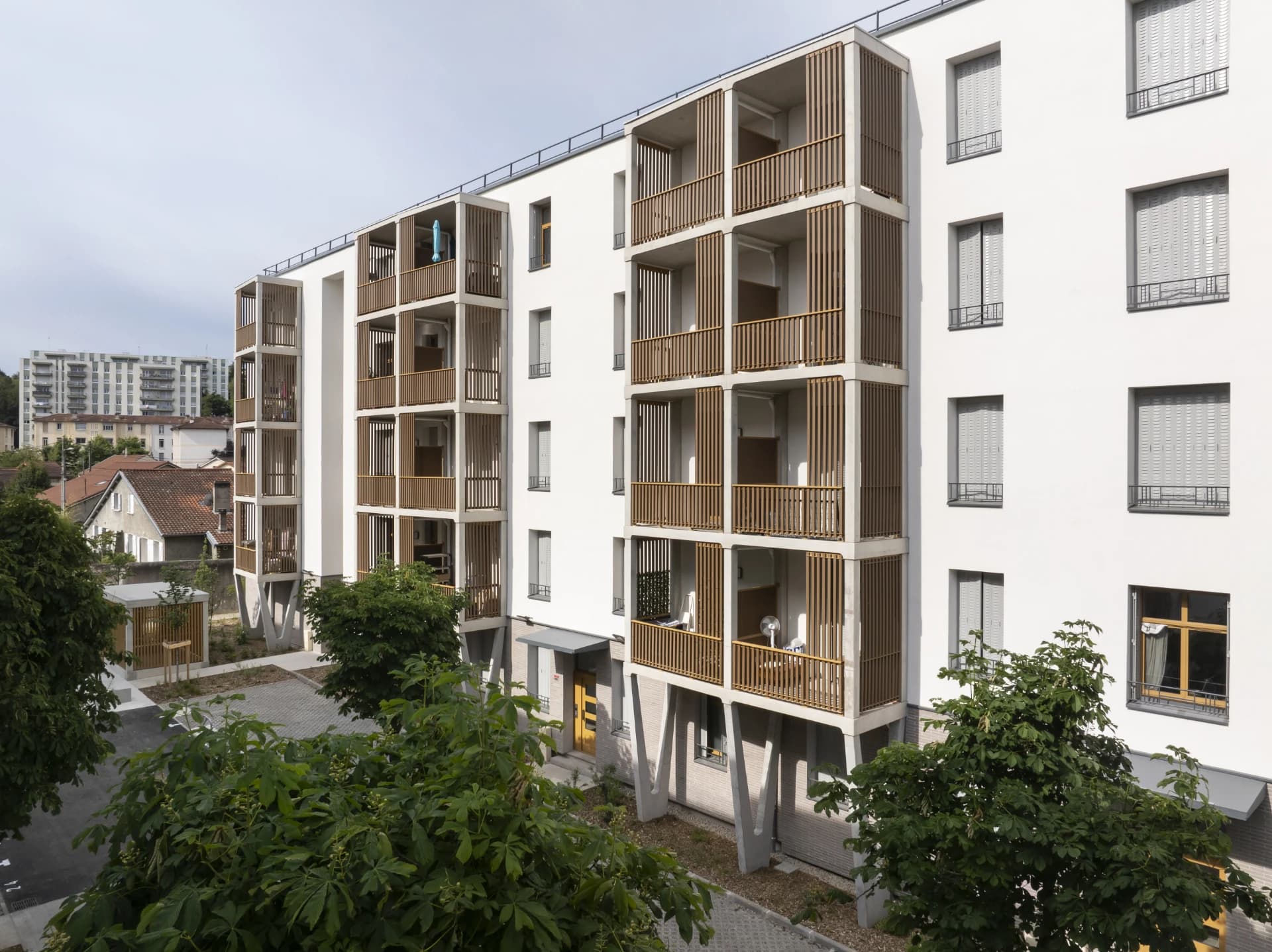 Façade d'un immeuble résidentiel moderne blanc à plusieurs étages, avec des balcons en béton étagés ornés de claustras et balustrades en bois clair, entouré de verdure sous un ciel lumineux.
