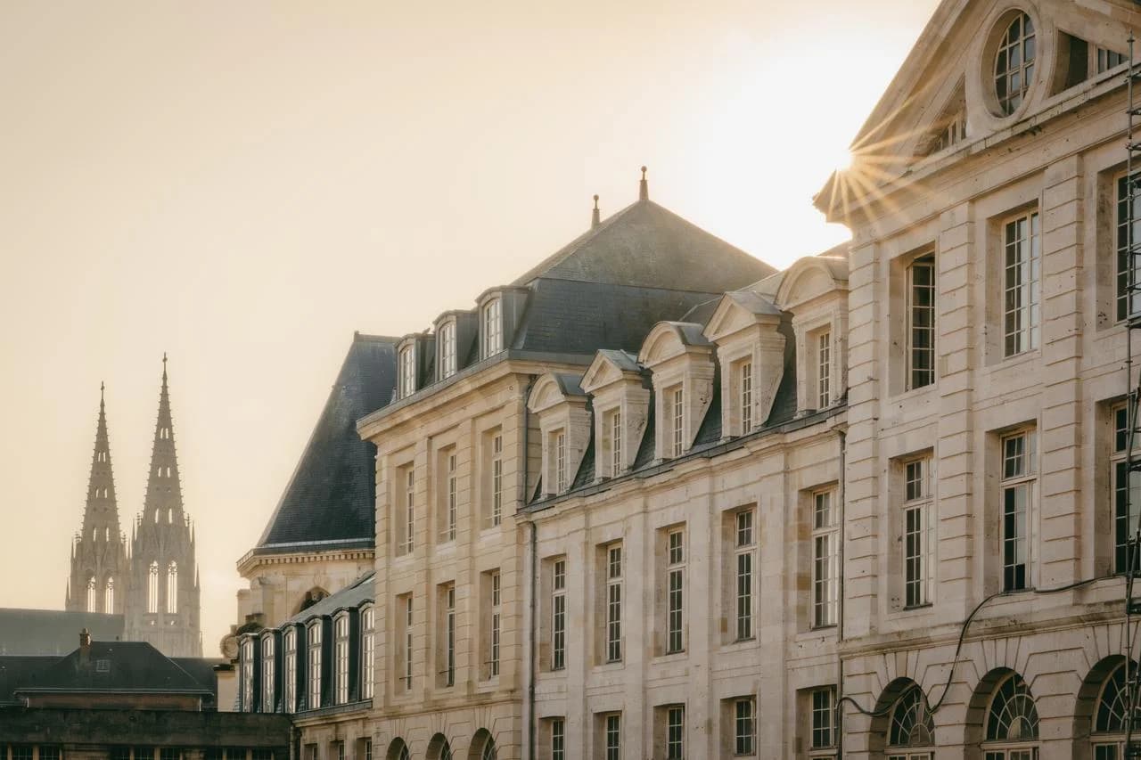 Façade de bâtiments anciens en pierre calcaire avec des toits en ardoise et de multiples fenêtres, éclairée par un soleil éclatant sur la droite, avec les flèches d'une cathédrale visibles au loin à gauche.