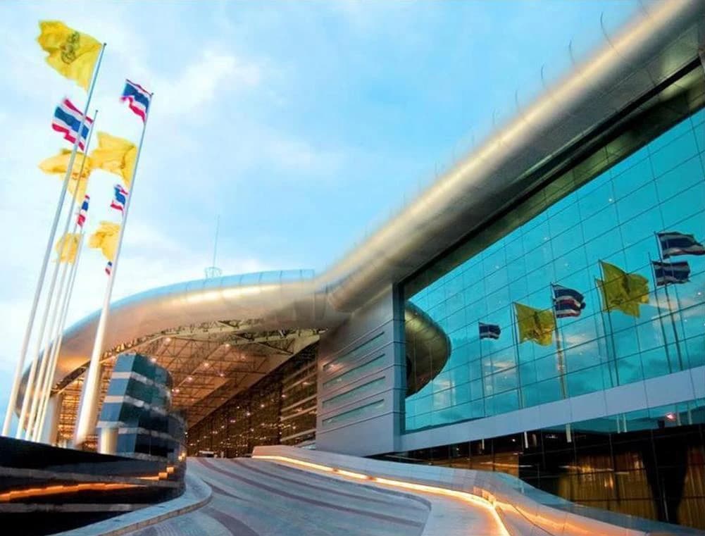Bâtiment moderne avec une façade en verre réfléchissante et de nombreux drapeaux thaïlandais et jaunes flottant devant, sous un ciel bleu.