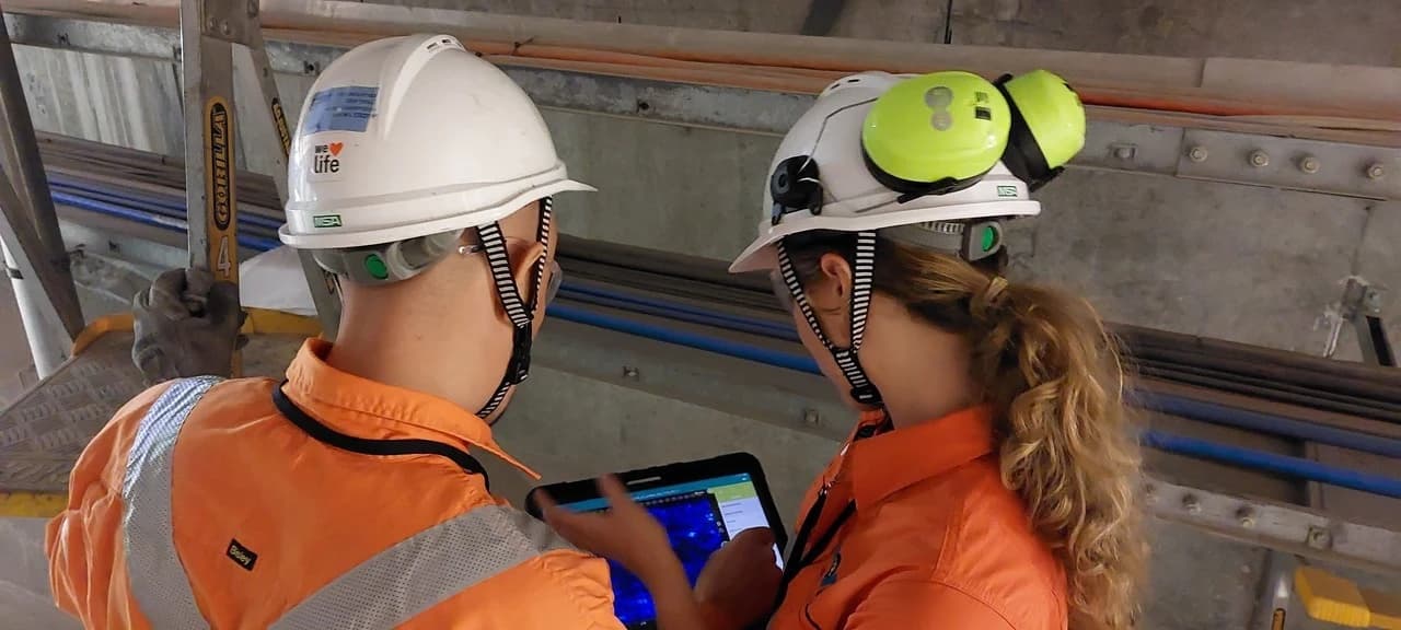 Vue aérienne d'un pont en construction au-dessus de l'eau, montrant une série de piliers en béton et de grandes structures d'assemblage métalliques bleues, avec des montagnes brumeuses à l'horizon.