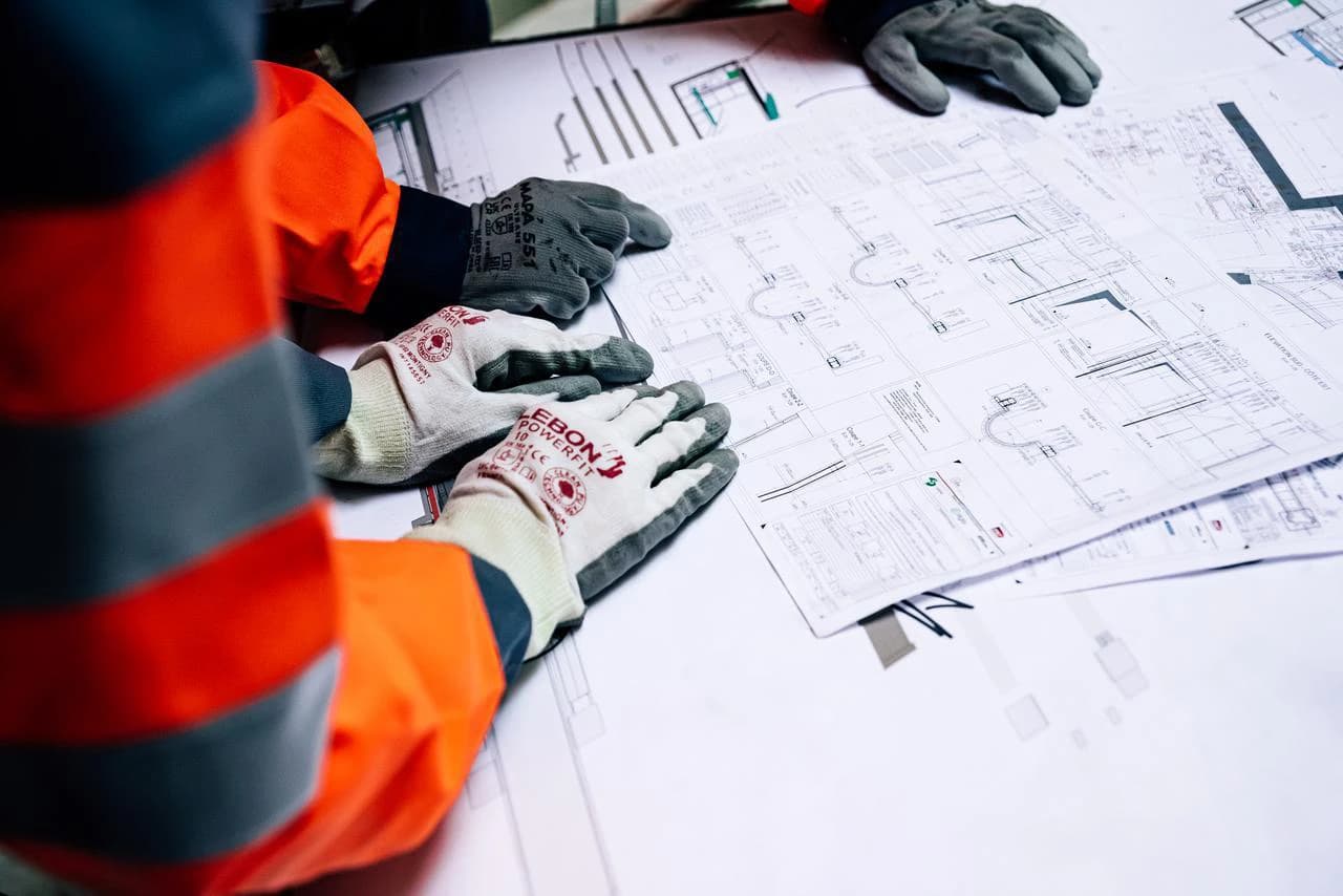 Mains gantées de deux personnes en vêtements de travail examinant des plans de construction ou des schémas techniques.