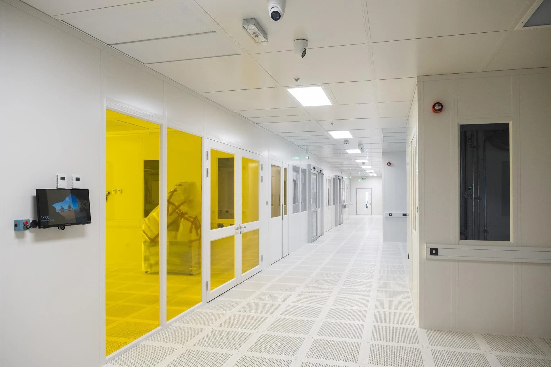 Long couloir de salle blanche stérile et lumineux, avec des murs blancs, un plancher surélevé perforé et des portes vitrées jaune vif menant à des zones de travail spécialisées, éclairé par des dalles lumineuses au plafond.