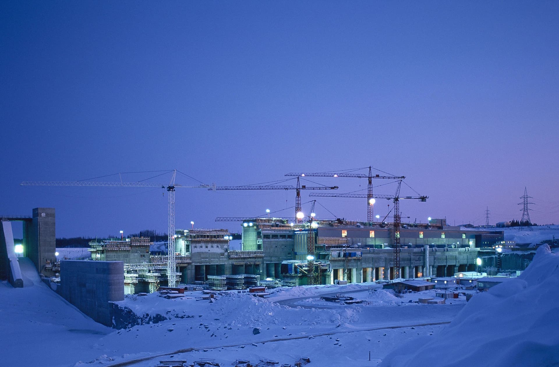 Vaste chantier de construction d'un barrage ou d'une centrale électrique la nuit, avec des structures en béton éclairées par des lumières vives et de multiples grues, sur un paysage enneigé sous un ciel bleu profond.