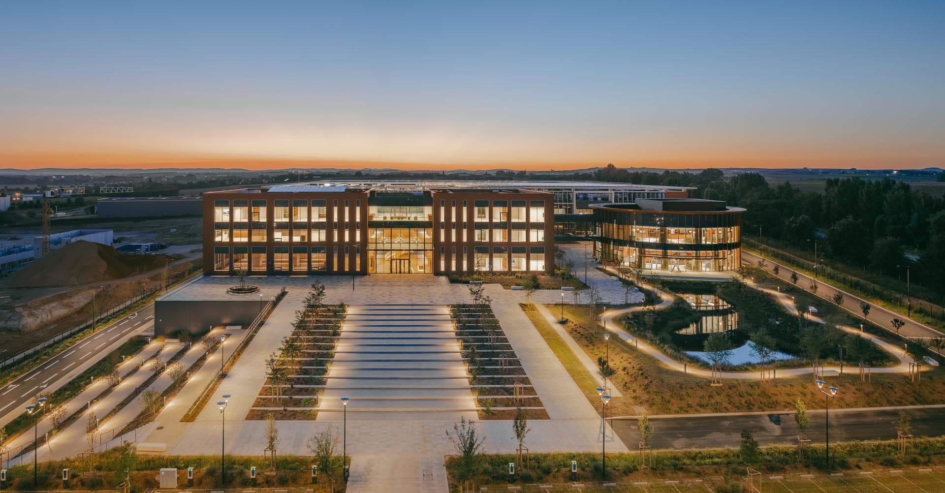 Vue aérienne d'un complexe de bureaux moderne au crépuscule. Les bâtiments aux façades marron clair et grandes baies vitrées sont illuminés, entourés d'une vaste place avec des chemins éclairés et un étang reflétant le ciel bleu et orange.