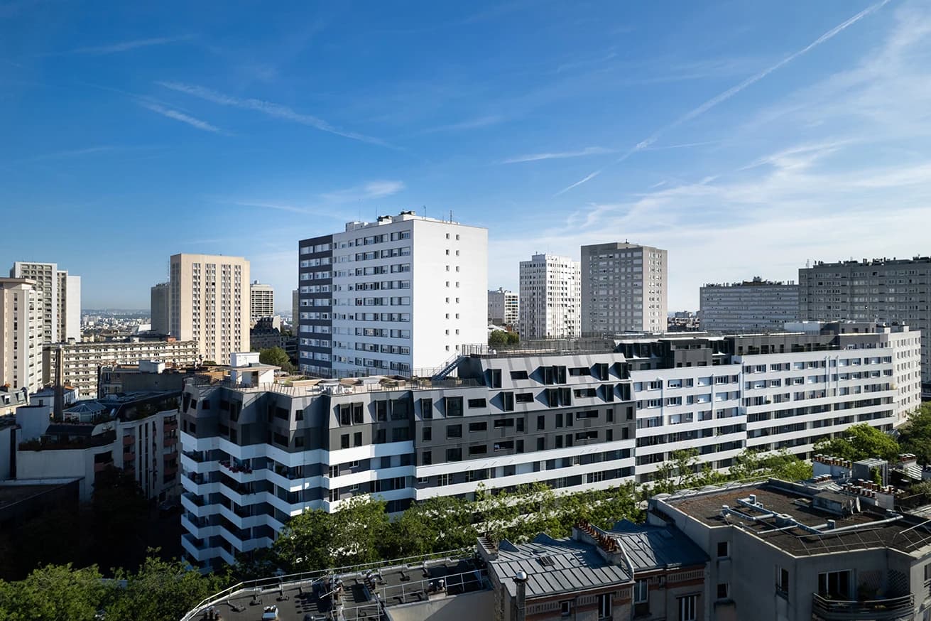Vue d'ensemble d'un paysage urbain dense avec des immeubles résidentiels variés, dont un à rayures noires et blanches et d'autres aux façades majoritairement blanches, grises et beiges, sous un ciel bleu clair. Des toits et des arbres verts sont visibles au premier plan.