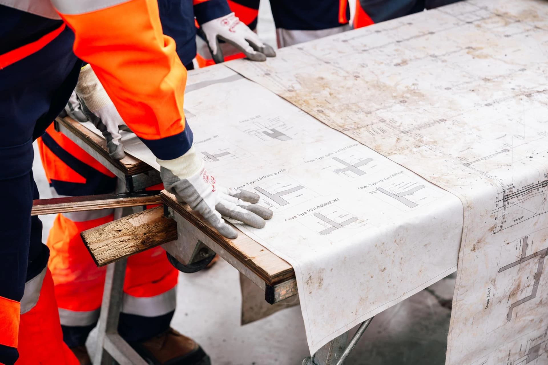 Des ouvriers en gants et tenues de sécurité orange et bleu marine examinent un grand plan de construction détaillé, légèrement sali, posé sur une surface en bois.