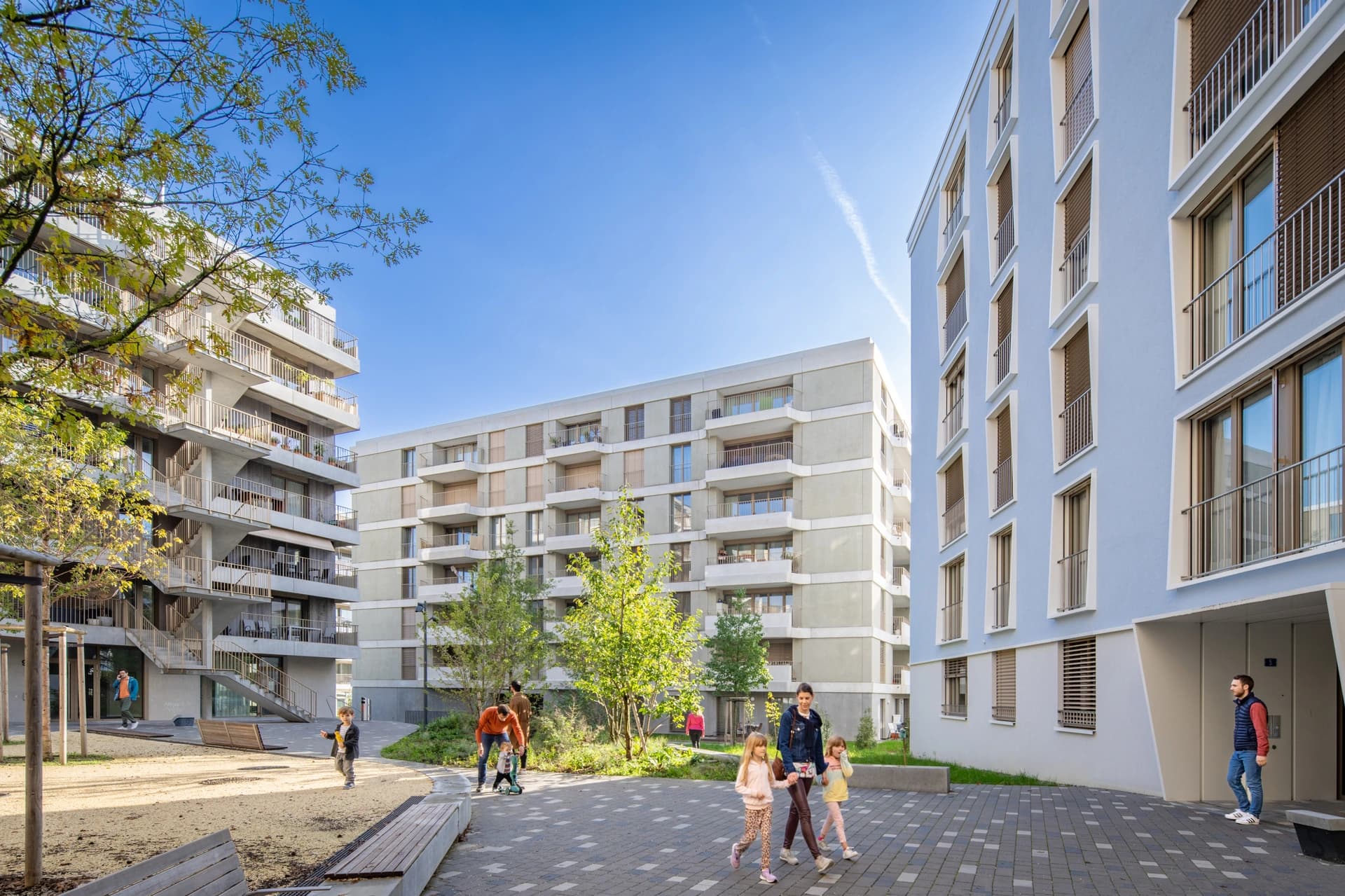 Modern apartment buildings in light blue and light grey surround a lively outdoor communal area with trees, paved paths, and a sandy play space where adults and children are enjoying a sunny day.