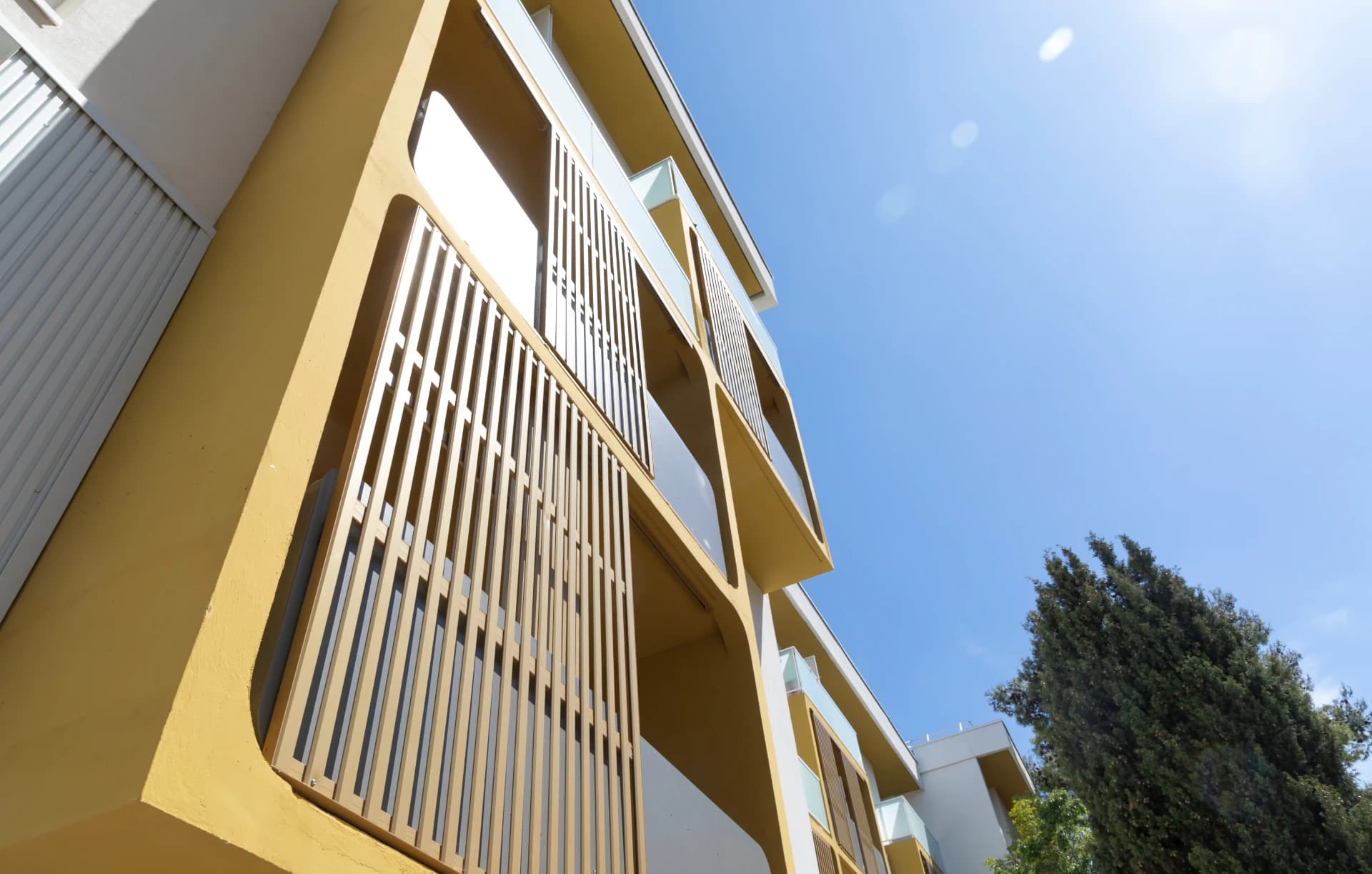 Façade angulaire d'un bâtiment moderne jaune-doré avec des balcons, des claustras à lattes verticales et des détails blancs, sous un ciel bleu éclatant avec un arbre vert visible à droite.