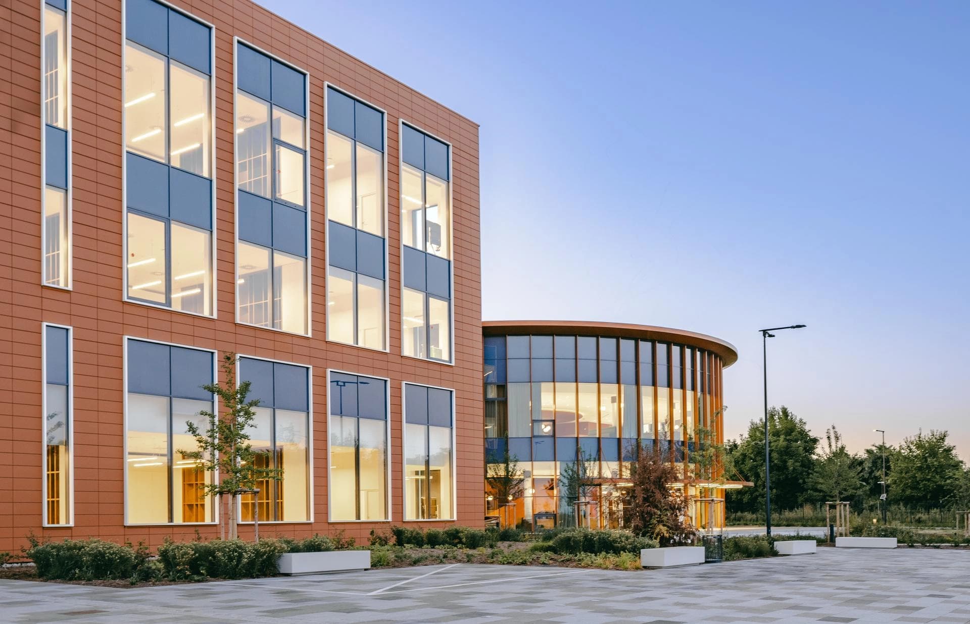 Deux bâtiments modernes et lumineux sous un ciel clair au crépuscule. L'un est rectangulaire avec une façade en carreaux brun-orange et de grandes fenêtres sombres, l'autre est rond avec une façade entièrement vitrée et des accents boisés, entourés d'un aménagement paysager.